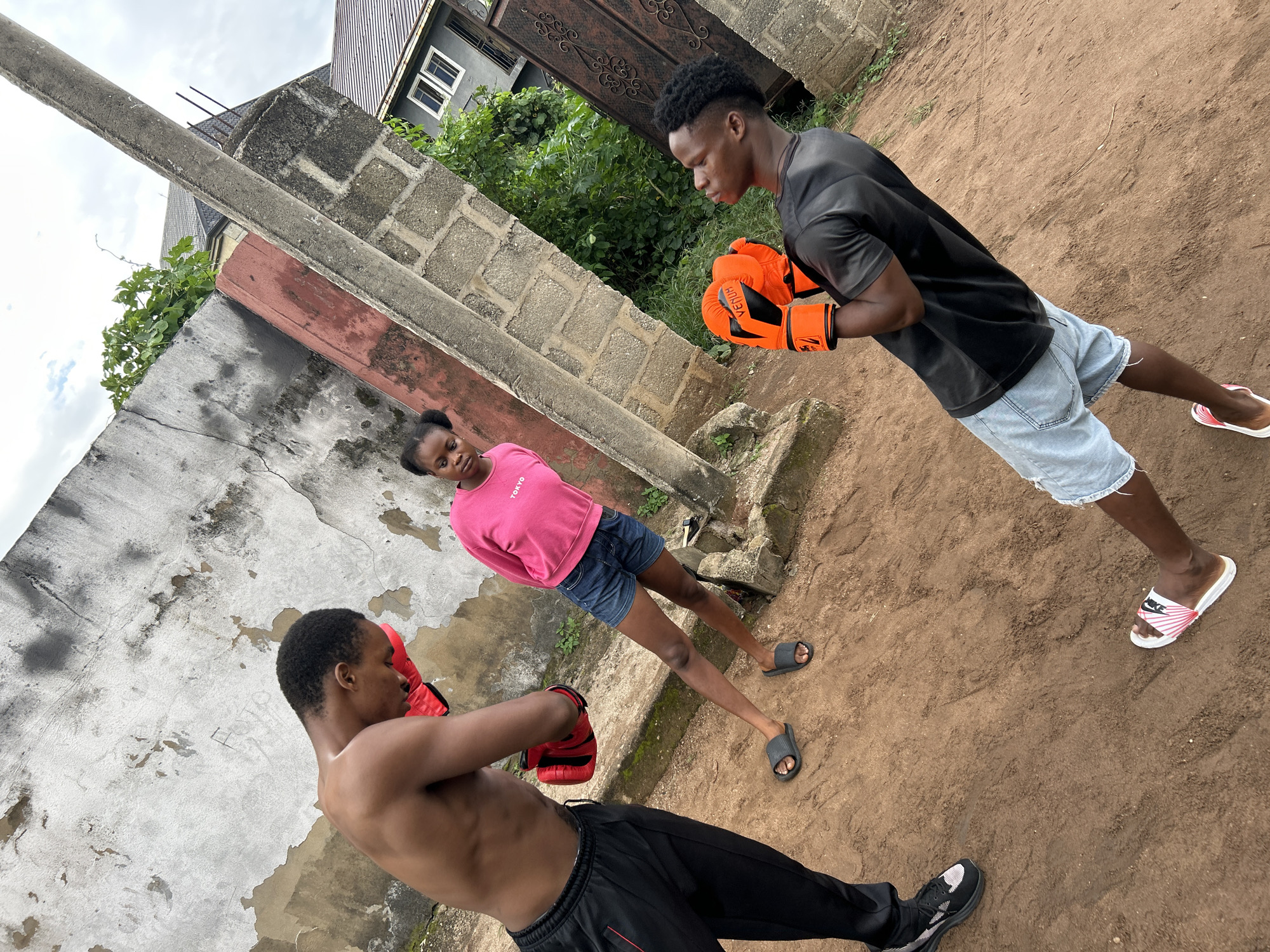 In Ihiagwa, Nigeria, three individuals are present in an outdoor environment characterized by dirt ground, rough concrete walls, and a cloudy sky. A shirtless man on the left, wearing black athletic pants and red boxing gloves, is positioned with his head bowed, appearing to be in a stance. To his right, a young woman in a pink long-sleeved shirt and denim shorts observes them. A second man on the right, wearing a black t-shirt and light denim shorts, also has red boxing gloves on and is facing the first man, his body angled and head turned slightly upwards. The background includes a weathered concrete wall with green vegetation growing on it, a tall concrete pillar, and a brick wall. The overall setting suggests an informal boxing practice or training session.