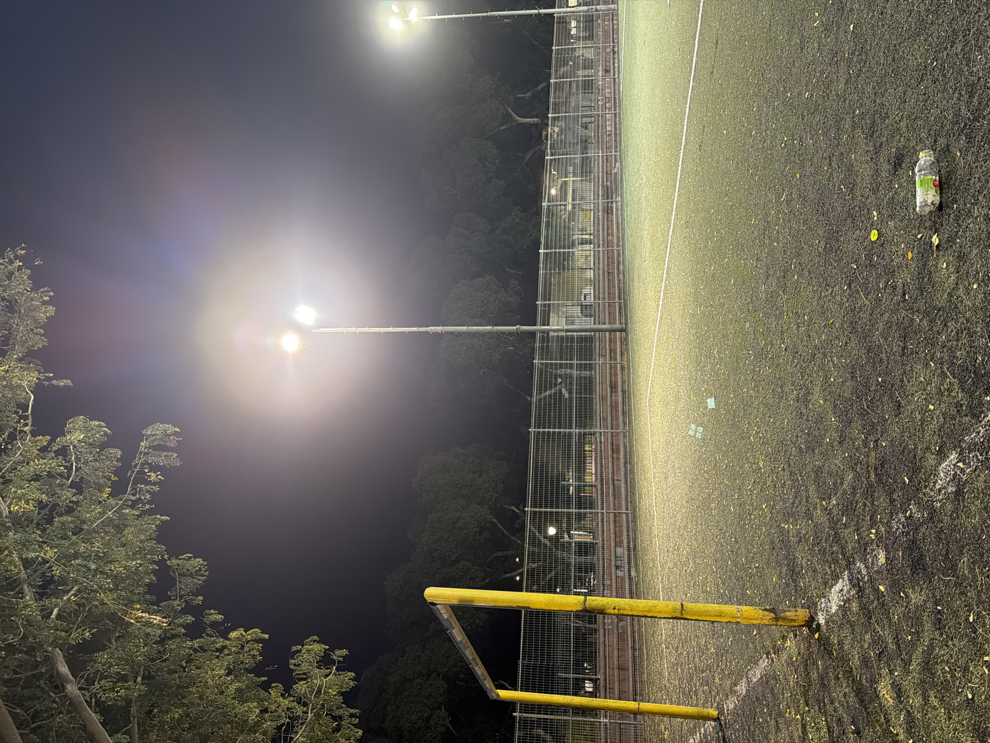 The image captures an artificial turf sports field at night, illuminated by several powerful floodlights in Kiryat Bialik, Israel. The dark sky indicates nighttime, with bright, almost blinding light sources creating significant lens flares and a diffused glow across the scene.
The foreground features a yellow metal goalpost on the lower left, extending onto the field. The ground is a dark green and black artificial turf, marked with distinct white lines that delineate the playing area. Numerous small, yellow leaf-like debris are scattered across the turf, particularly noticeable in the illuminated areas. Towards the upper right of the field, a discarded, crumpled plastic water bottle, predominantly white with some green elements and faint, unreadable text, lies on the ground. Two small, light-colored pieces of litter are also visible on the field near the center.
A tall, chain-link or wire mesh fence runs along the right side of the field, separating it from a darker, wooded area where trees are dimly visible. Additional trees and dense foliage are present in the lower left corner of the frame, silhouetted against the artificial light. No people or specific activity are visible, suggesting the field is empty, perhaps after a game or awaiting future use. The scene conveys a sense of stillness and artificial illumination contrasting with the natural darkness of the night.
