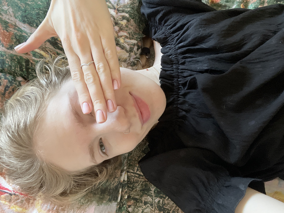 A woman with blonde hair lies on her back, partially obscuring her face with her hand. She has light skin, a silver nose ring, and wears a silver ring on her hand. She gazes towards the camera with one visible eye. She is wearing a black, gathered-neck top. The background shows a patterned surface of green and brown tones. The image is rotated slightly, with the woman's face angled towards the left side.