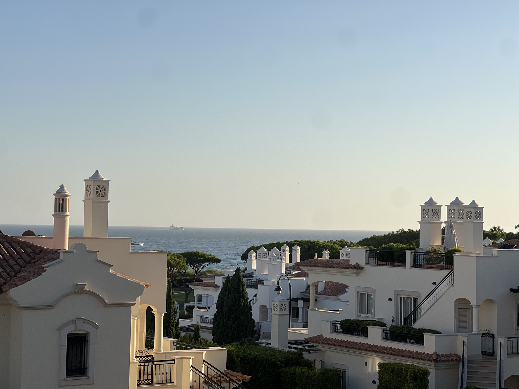 The image depicts a view of white buildings with terracotta roofs, characteristic of the architecture found in Almancil, Portugal. The structures appear to be modern residences or resort properties, with multiple levels, balconies, and decorative chimneys. Lush green trees and manicured bushes surround the buildings, suggesting a well-maintained landscape. In the background, the calm, blue sea stretches to the horizon, with a large ship visible in the distance. The sky is a clear, pale blue, indicating fair weather. The scene is bathed in warm, soft light, suggesting it was captured during either sunrise or sunset. There are no people visible in the image.
