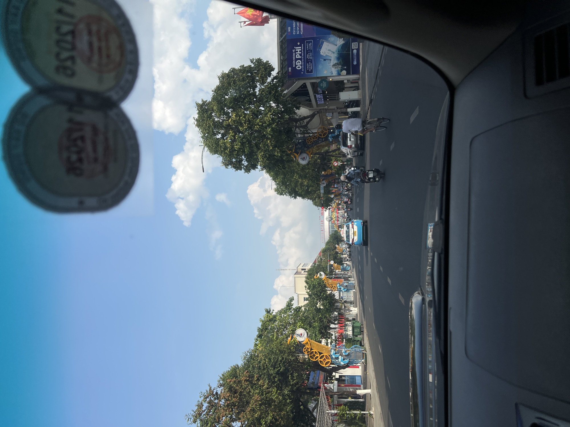 This media file captures a bright daytime street scene in Nha Trang Ward, Vietnam, viewed from inside a vehicle. The image is oriented vertically (originally rotated 90 degrees clockwise).
The scene depicts a busy urban street with a road, buildings, and lush green trees lining both sides. Numerous vehicles are visible, including motorcycles, bicycles, and at least one car (a blue taxi). Several people are present, cycling or riding motorbikes, indicating daily commute or travel.
Notable details include a clear blue sky with scattered white clouds, suggesting good weather. In the foreground, two blurred, circular objects, possibly bottle caps or similar items, are visible with the mirrored text "ASSOSE" on them. Along the street, decorative elements resembling large yellow and blue mechanical structures or stylized objects are placed between trees. On a building to the left, Vietnamese flags are flying, and a large billboard advertises "MB" (likely a bank), featuring text such as "NHẬN NGOẠI TỆ" (Receive Foreign Currency), "CỨ 2 NGƯ," "SỬ DỤNG" (Use), "ĐĂNG KÍ NGAY" (Register Now), and prominently "O Đ PHÍ" (likely "0 Đồng Phí" or No Fee). The perspective from inside a car is evident from the visible dashboard and windshield framing the scene.
