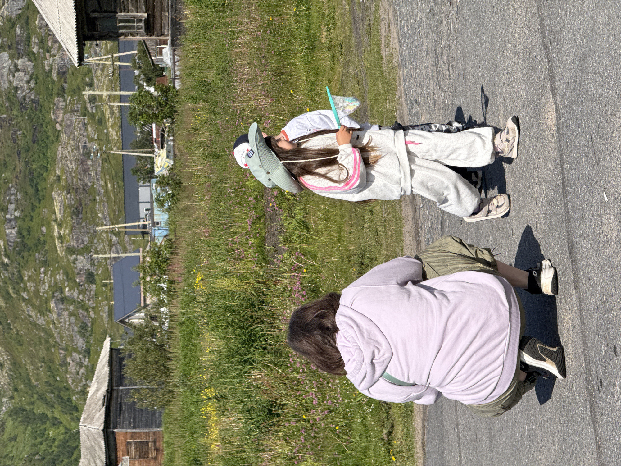 In Teriberka, Russia, a person wearing a light purple hoodie and olive green cargo pants crouches on an asphalt road. The person is viewed from behind, with their head turned slightly to the right. Two children walk in front of the person on the road. The child on the left wears a large, light green wide-brimmed hat and a light gray tracksuit with pink stripes. This child holds a blue butterfly net. The child on the right, partially obscured, wears a white shirt and a baseball cap. In the background, there is a grassy area with scattered pink and yellow wildflowers. Beyond the grass, several wooden buildings with weathered roofs are visible. Further back, a rocky, green hillside rises. Power lines and poles are also present in the mid-ground. The weather appears sunny.