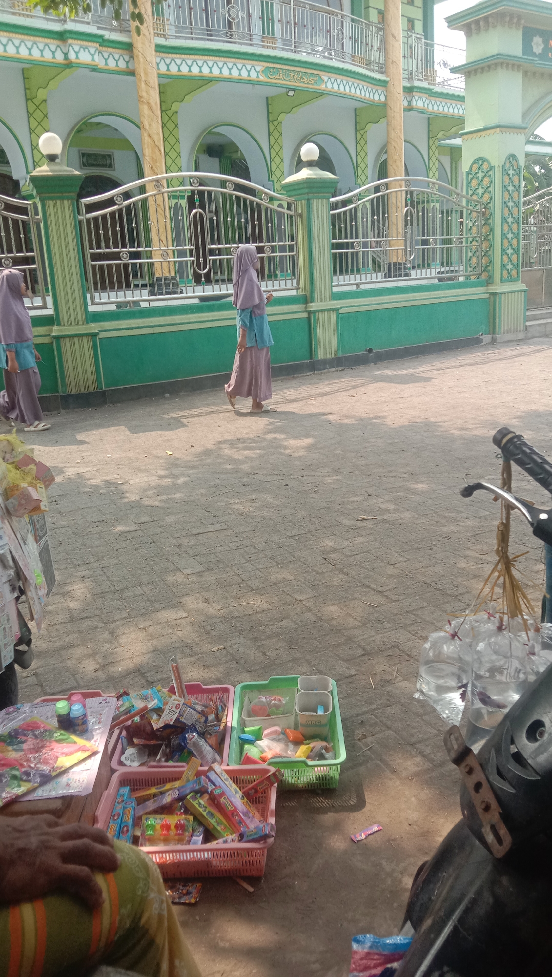 In the city of Ngempit, Indonesia, a street vendor displays an assortment of goods, including small toys, candies, and bottled water, arranged in colorful baskets on the ground. Two young girls in hijabs walk past a mosque in the background, its green and white facade adorned with intricate architectural details. The ground is paved with textured tiles, and the scene is bathed in the warm glow of sunlight, casting long shadows across the pavement. A motorcycle is partially visible in the foreground, with its handlebars and a bundle of bottled water hanging from it. The overall atmosphere is one of daily life unfolding in a vibrant community.