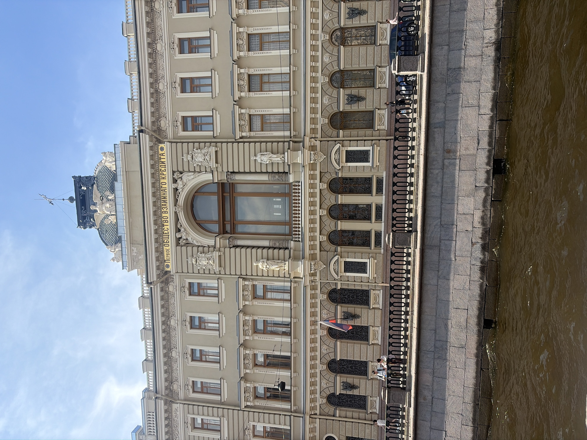 This is a full-shot, eye-level, exterior photograph of the "First Mutual Credit Society" building located in Saint Petersburg, Russia. The building is a grand, light beige edifice with ornate architectural details, including decorative moldings, statues, and numerous arched windows. The inscription "ПЕРВОЕ ОБЩЕСТВО ВЗАИМНАГО КРЕДИТА" (First Mutual Credit Society) is visible in gold lettering above the central arched entrance. A Russian flag is displayed on a flagpole in front of one of the arched windows on the lower level.
In the foreground, a canal with brown water runs along the bottom of the image, bordered by a granite embankment. On the embankment, a black ornate railing separates the walkway from the canal. Several people are visible on the walkway, including two individuals standing and conversing, a woman walking, and a child. A dark blue SUV and a dark-colored car are parked on the walkway. The sky above is a clear blue with some wispy white clouds. The overall scene depicts a sunny day in Saint Petersburg, showcasing the historic architecture of the city.