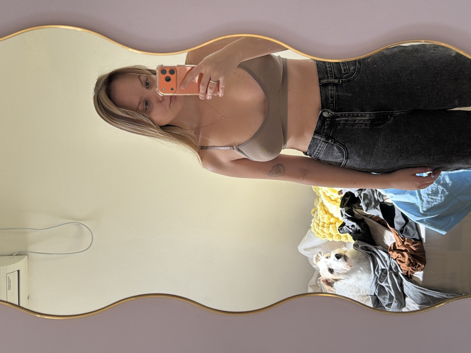 The image displays an indoor scene captured via a mirror selfie. A female subject with light brown hair, wearing a light brown brassiere and dark grey denim jeans, is reflected in a wavy, gold-framed mirror. She holds an orange-cased smartphone in her right hand, positioned to capture the image. A faint tattoo is visible on her left bicep, and a ring is present on her right ring finger.
Behind her reflection, a white-and-brown terrier-type dog is situated on a bed or sofa, which is partially covered with assorted clothing items in shades of grey, brown, and blue, alongside a yellow textured cushion. A white split-system air conditioning unit is mounted on the pale yellow wall in the upper left background. The scene is consistent with a residential interior. There are no discernible elements within the image that confirm the location as Tel-Aviv, Israel.