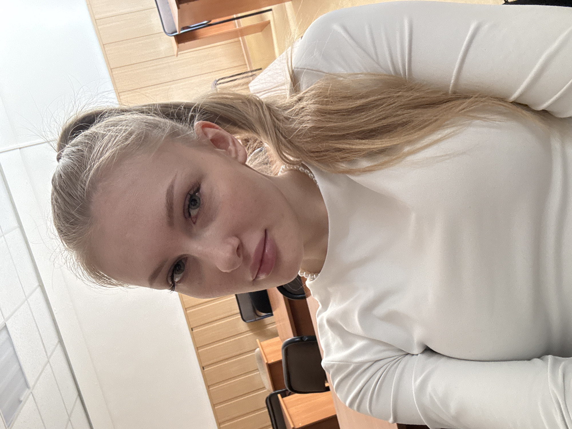 The image, taken in Perm, Russia, features a young woman captured in a rotated, close-up indoor portrait. The woman has light blonde or light brown hair, pulled back into a ponytail, with some softer strands framing her face. She has light blue or green eyes and a calm, pleasant expression with a subtle hint of a smile. Her lips are a natural pink. She is wearing a plain, long-sleeved white top and a white pearl necklace.
The background suggests an indoor, institutional setting, possibly a classroom or lecture hall. Light-colored wooden wall paneling is visible, along with glimpses of dark chairs and wooden desks arranged in what appears to be rows. The lighting is soft and even, typical of interior spaces. There is no visible text in the image. The overall impression is a candid or selfie-style portrait taken in a structured environment.