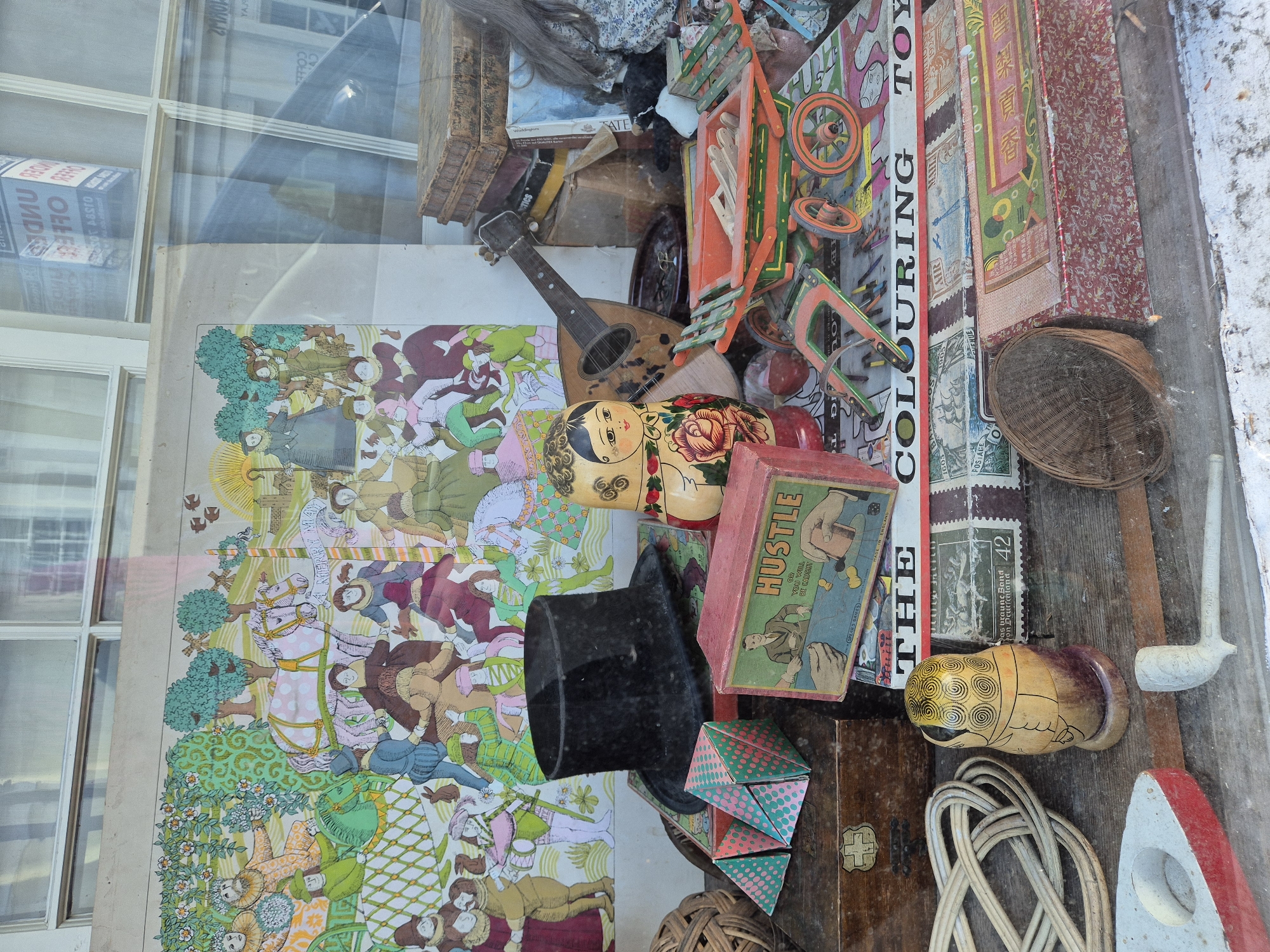 The image shows a window display in what appears to be a vintage shop, probably in West Suffolk, United Kingdom. A variety of antique toys and collectibles are densely arranged, including a large illustrated poster depicting figures from a historical or fairy tale setting. Dominating the display are a nesting doll (matryoshka), a mandolin or similar stringed instrument, a child's wagon toy, and various boxes and containers. Visible is a box of the game "Hustle," a box labeled "The Colouring Toy," and other items of uncertain origin. Stamps and vintage-themed decorations add to the eclectic collection. The window reflection reveals a partial view of buildings across the street. The general aesthetic is cluttered and nostalgic.