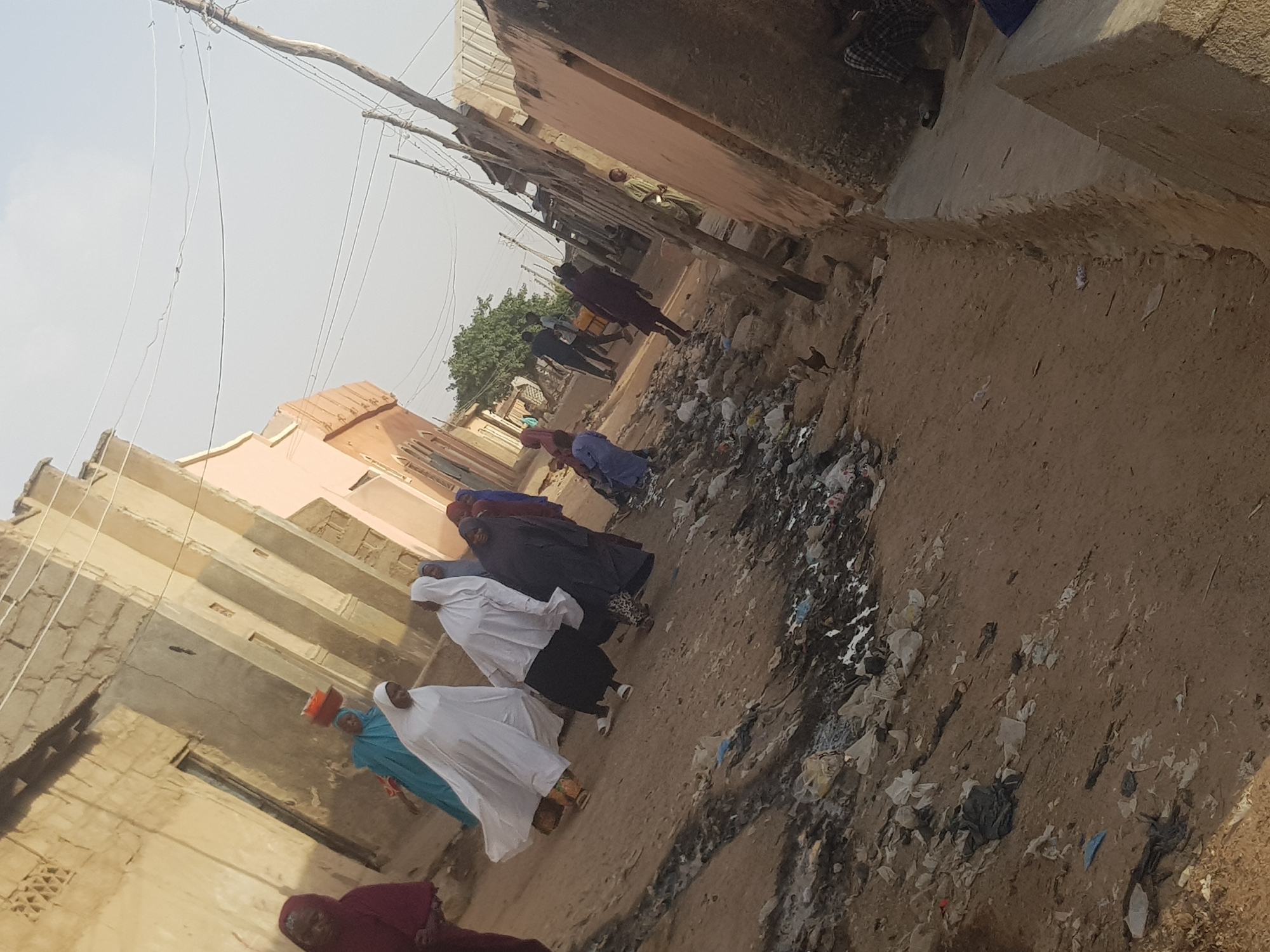 A street in Dutsinma B, Nigeria, displays trash accumulation on its sandy surface. A group of individuals dressed in traditional attire are walking along the street. Buildings, some with damaged walls, line both sides of the narrow passage. Overhead, power lines are visible.