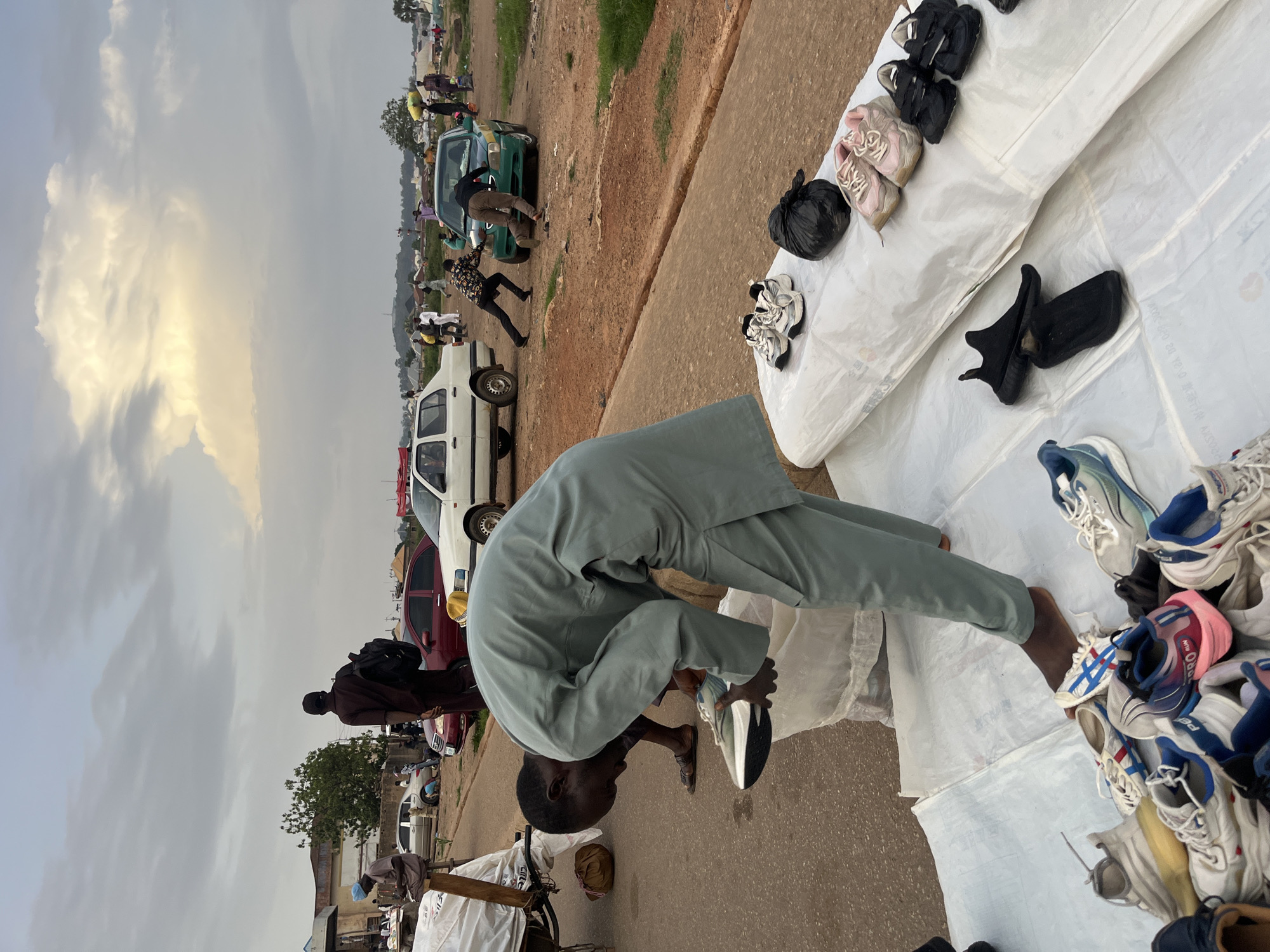 The image depicts an outdoor market scene in Bukuru, Nigeria. In the foreground, a man wearing a light green traditional outfit is bent over, holding a white sneaker with blue and grey accents in his right hand. He appears to be sorting through a large white bag or container of shoes. In front of him, spread on a white tarp, is a collection of various sneakers, including white, blue, pink, and black pairs, some with intricate designs.
In the mid-ground, several cars are parked on a paved road. A green taxi is being pushed by two men. To the left, a red car and a white sedan are visible. A few other people are scattered around, some standing and observing, while others appear to be engaged in their activities. Further in the background, a settlement with buildings and trees is visible under a cloudy sky with hints of sunlight breaking through. The ground is a mix of paved road and reddish earth. The overall impression is of a busy, informal marketplace.