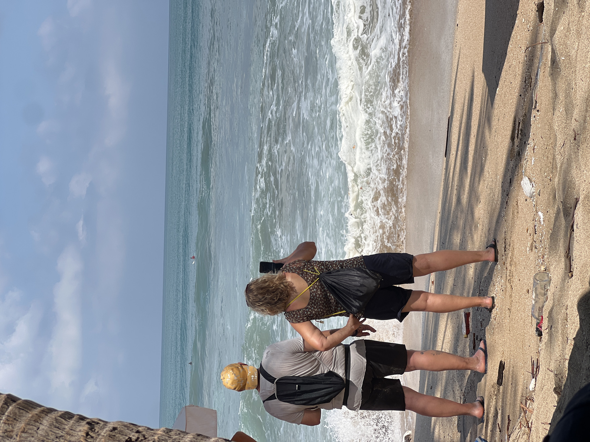 In Kuta, Indonesia, two individuals stand on a sandy beach, facing the ocean. The person on the left, wearing a yellow cap, a grey t-shirt, and black shorts, has a black backpack on. The person on the right, with blonde curly hair and wearing a patterned top and black shorts, is holding a black smartphone and appears to be taking a picture. The ocean has gentle waves and a few surfers can be seen in the distance. The beach is littered with debris, including plastic bottles and driftwood. The sky is partly cloudy with a bright, hazy sun.