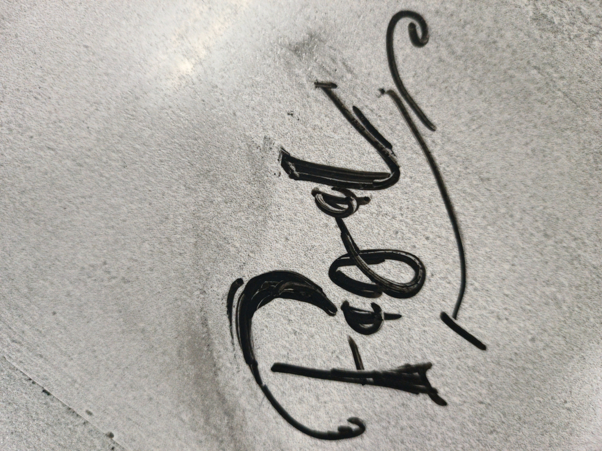 The image displays a close-up, slightly angled view of black ink calligraphy on a textured, light-grey surface. The calligraphy appears to be a signature or a short word rendered in a flowing, cursive style. The ink is thick and somewhat irregular, with some areas appearing more opaque than others, suggesting it was applied with a brush or similar tool. The background surface is speckled with darker grey and white particles, giving it a granular or rough texture, reminiscent of concrete or a textured paper. There are no discernible elements of the city of Ngoro, Indonesia, present in the image. The lighting is somewhat uneven, with a bright, possibly reflective area visible in the upper left portion of the image, casting subtle shadows. No people or specific objects other than the calligraphy and the surface are visible.