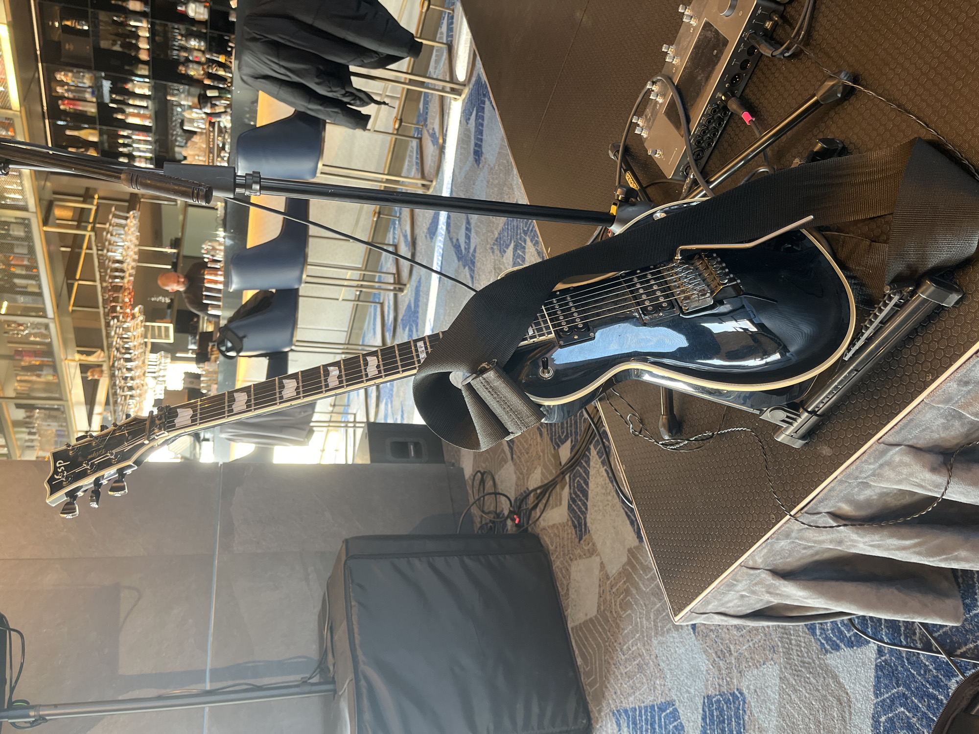 This image, captured in Sofia, Bulgaria, depicts a music performance setup within a bar or lounge environment. The main subject is a black ESP LTD Eclipse electric guitar, prominently displayed and resting on what appears to be an amplifier or a stand. The guitar features a white binding along its body and neck, six strings, and distinctive block inlays on the fretboard. A black fabric strap is draped over its body, and a tremolo system is visible near the bridge. The headstock clearly displays the brand "ESP LTD" and the model "Eclipse."
Adjacent to the guitar, on a raised platform, sits a complex piece of music equipment, likely a multi-effects processor or audio interface, characterized by an array of knobs, buttons, and various input/output jacks, suggesting a professional setup. Cables are seen connecting different components, and a microphone stand extends vertically from the foreground. On the patterned carpet below, which features geometric designs in blue, brown, and off-white, a black soft case or bag lies near additional cabling.
The background reveals a well-appointed bar area. Shelves are densely packed with numerous bottles of alcohol and glassware, suggesting a wide selection. Several bar stools with dark blue seats are visible in front of the bar counter. A person in a dark top is faintly discernible behind the bar, their face and activity indistinct. The walls appear to be constructed from a grey, possibly concrete or stone, material. Bright ambient lighting suggests the scene is taking place during the day or in a very well-lit interior. The overall impression is a venue prepared for a live musical performance.