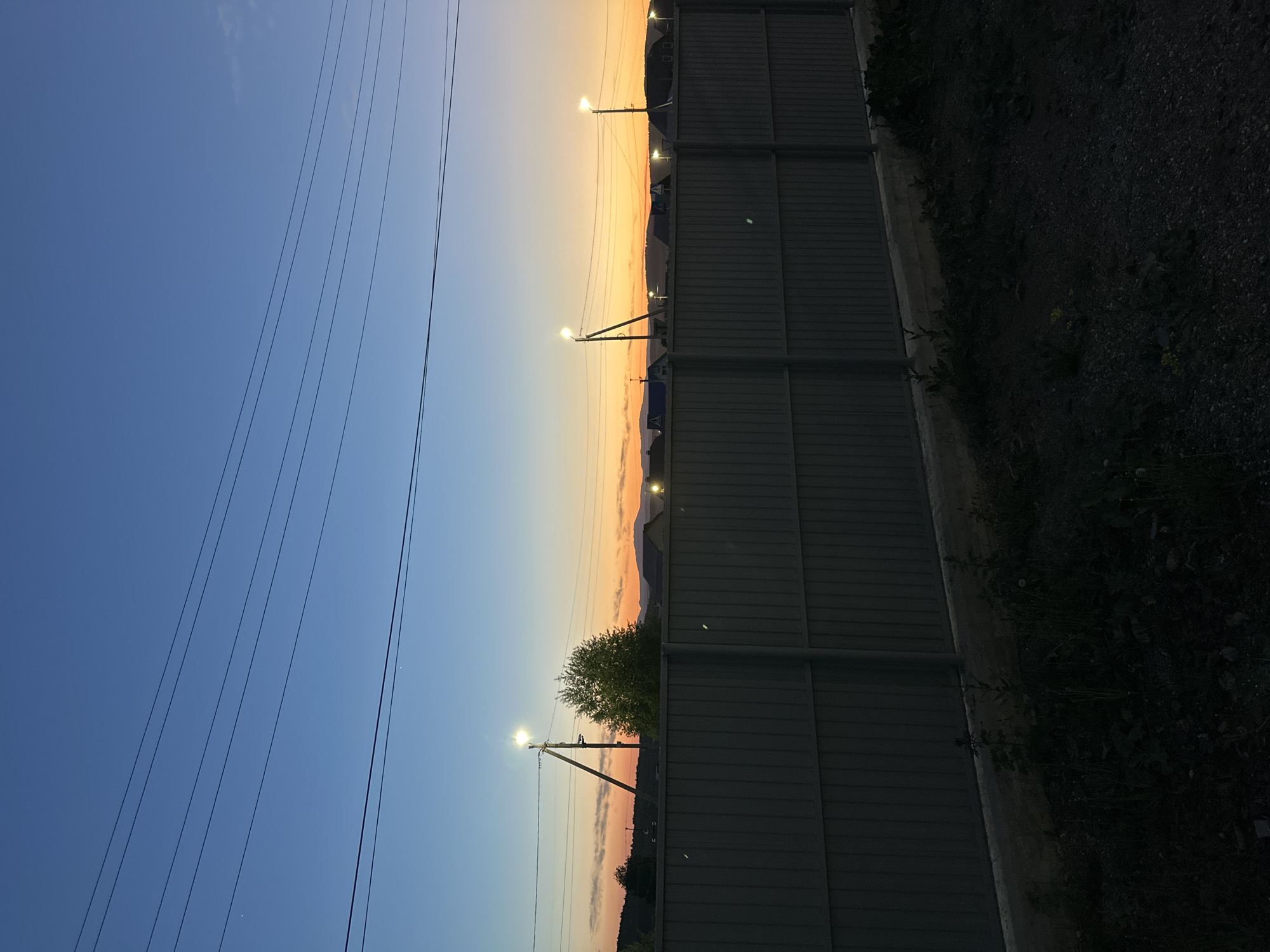 The image shows a scene at dusk in an urban area. On the left, power lines stretch across a gradient sky that transitions from blue to orange near the horizon. A series of streetlights are illuminated along the horizon, positioned between a dark metal fence on the right and a distant, low-lying landscape with vegetation. The ground to the right of the fence appears to be a mix of dirt and gravel, partially obscured by shadow. A tree is visible amidst the streetlights.