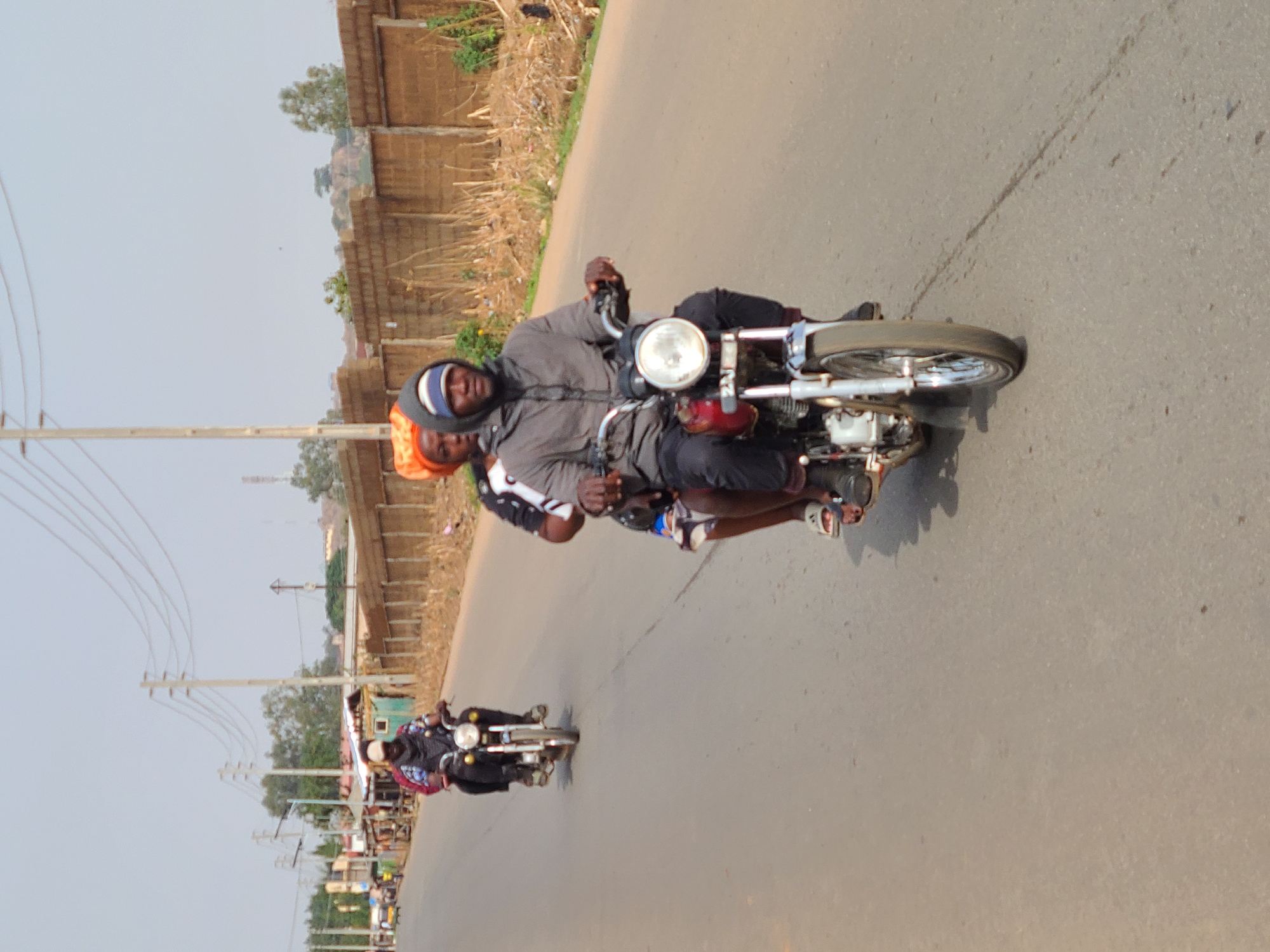 An outdoor, eye-level shot captures several individuals riding motorcycles on a paved road, likely in Barakin Lerawi, Nigeria. In the foreground, two men are on a motorcycle. The driver wears a grey hooded jacket over a light-colored head covering, black trousers, and shoes, with his hands on the handlebars. A passenger, partially obscured behind him, wears an orange head covering and a white shirt with dark patterns. Both appear focused on the road.
Further down the road, a second motorcycle is visible with at least one rider, also in dark attire. The road surface shows some tire marks and debris. To the right of the road, the shoulder is lined with dry vegetation, some refuse, and various structures, including what appear to be unplastered brick buildings and fences. On the left side of the road, multiple utility poles with overhead wires run parallel to the street, with more distant buildings and trees visible. The sky is bright and clear, indicating daylight hours with fair weather. There is no visible text in the image.