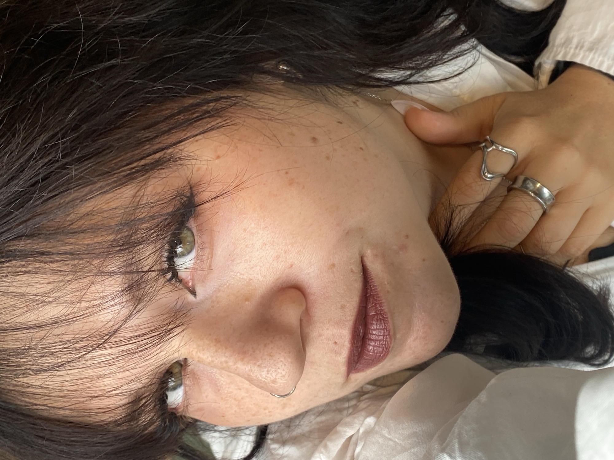 This is a close-up, self-portrait-style photograph of a young woman with dark hair and fair skin, lightly dotted with freckles. She has green eyes with visible eyeliner and mascara, and her lips are painted with a dark, matte lipstick. A silver hoop nose ring is visible in her left nostril. She is wearing a white collared shirt. Her right hand is touching her neck, and she is wearing two silver rings: one a simple band on her ring finger, and another larger, more elaborate ring shaped like an abstract heart or open loop on her index finger. The background is soft and out of focus, suggesting an indoor setting. There is no visible information in the image that would indicate a specific location in Saint Petersburg, Russia.