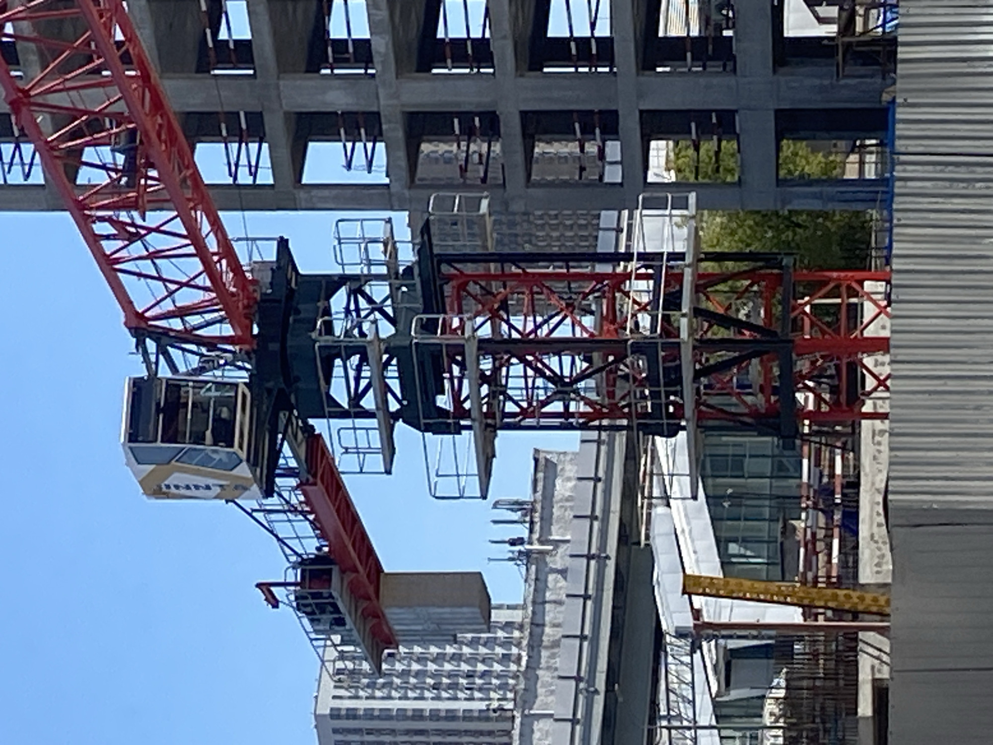 The image shows a construction site in Moscow, Russia, dominated by a large tower crane. The crane has a red boom and a white and gold operator's cab. In the background, several high-rise buildings are visible, some of which appear to be under construction as well. The foreground shows a concrete structure with visible rebar, indicating ongoing construction work. A metal fence runs across the bottom of the image, further enclosing the site. The sky is clear and blue, suggesting fair weather.