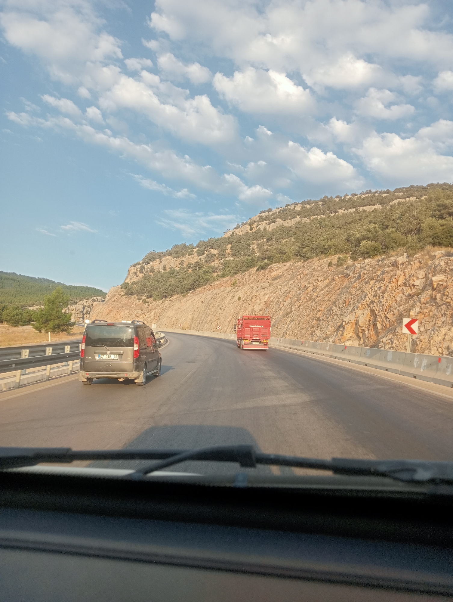An eye-level, medium shot captures a view from inside a vehicle looking out at a highway. The highway curves to the right, flanked by a rocky, vegetated hillside on the left and a concrete barrier with guardrails on the right. A dark-colored minivan is visible on the highway ahead of the viewer's vehicle, with a license plate that reads "07 DAE 16." Further down the road, a red truck is also traveling in the same direction. The sky above is a clear blue with scattered white clouds, and the sun appears to be setting, casting long shadows. The scene is set in a mountainous, rural area, likely on the outskirts of Korkuteli, Turkey, given the license plate.