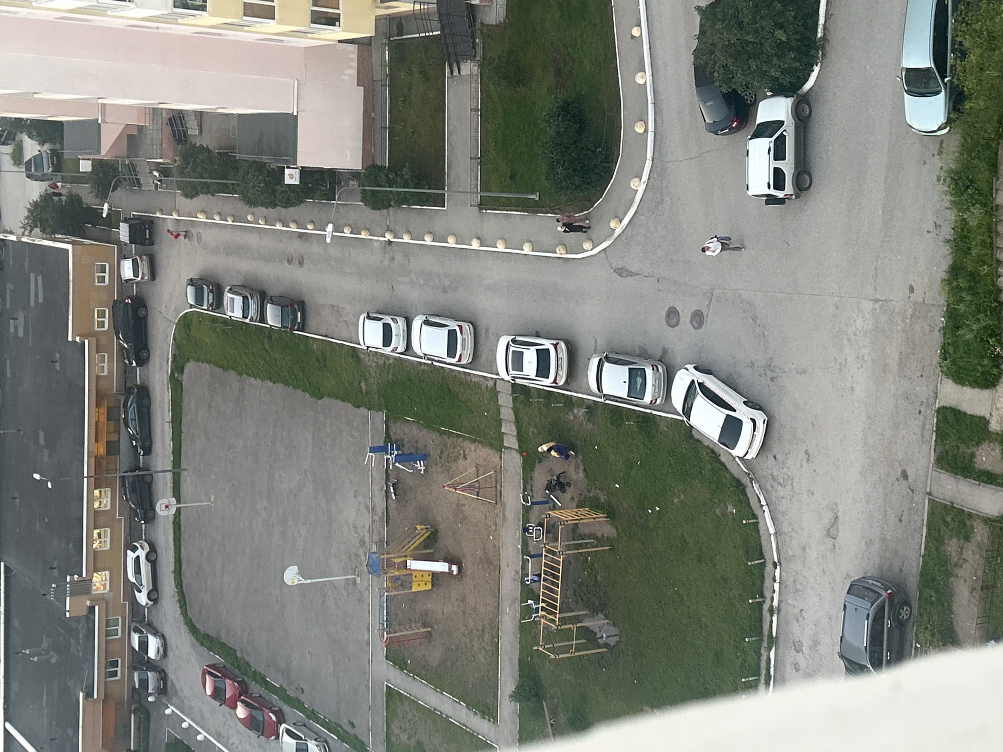 The image depicts an aerial view of a residential courtyard in Perm, Russia, likely during daytime or early evening. The scene is dominated by a multi-story residential building on the right, partially visible, with a pale pink facade and yellow accents. A grey asphalt road or parking area winds through the courtyard, lined with white spherical bollards.
Several vehicles are parked along the road. These include a light blue sedan, a dark grey SUV, a white SUV with a spare tire on the back, a black sedan, and a row of white sedans further up the road. There are also two red sedans parked in a lot to the upper left of the image, near a playground.
The courtyard features green grassy areas interspersed with paved sections. A playground with slides, swings, and climbing structures is visible in the lower left quadrant. Another area with outdoor exercise equipment is also present.
Two individuals are walking along the paved pathway in the lower right section of the image. Another person is walking a short distance away, carrying a bag. In the middle ground, near the playground, two people are standing, one of whom appears to be a woman with a stroller.
The overall impression is a typical urban residential area with parking, recreational facilities, and pedestrian activity.