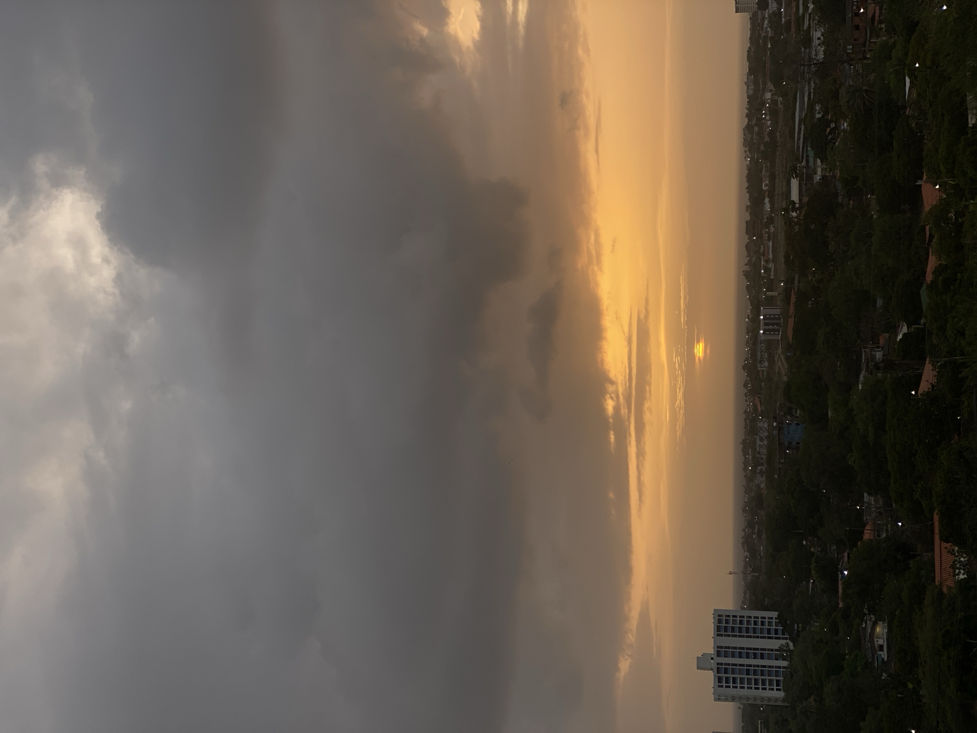 This image captures a cityscape of Juan Griego, Venezuela, during a dramatic sunset. The sky is dominated by heavy, dark grey clouds that transition into a vibrant orange hue near the horizon, where the sun is partially obscured. A sliver of the sun is visible, casting a warm glow. Below the sky, a densely treed landscape is interspersed with urban structures, including several buildings. Prominently featured on the left is a tall, white multi-story building with a dark-framed window pattern. In the distance, a hazy ocean or large body of water can be seen. The overall atmosphere is moody and atmospheric, typical of a pre-storm or post-storm twilight.