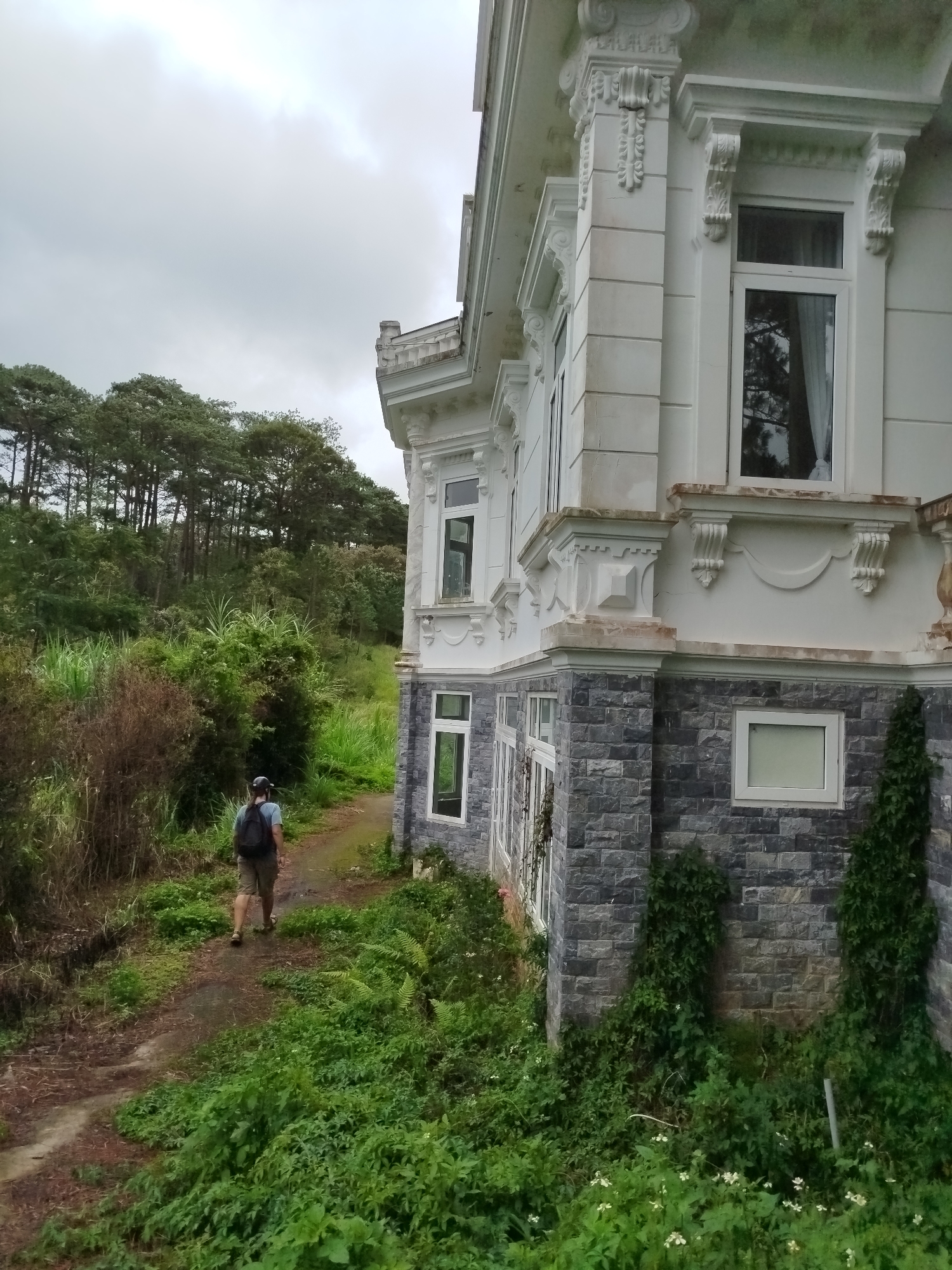 A person with a backpack walks away from the camera on a dirt path that is partially covered by vegetation. The path leads alongside a large, ornate white building with dark stone accents. The building has multiple windows and intricate decorative architectural details. Lush green foliage, including trees and tall grass, surrounds the path and the building. In the background, a dense forest of tall pine trees covers a hillside under a cloudy sky. The overall scene suggests an outdoor setting, possibly on the outskirts of Da Lat, Vietnam, known for its hilly terrain and pine forests, and the presence of European-style architecture.
