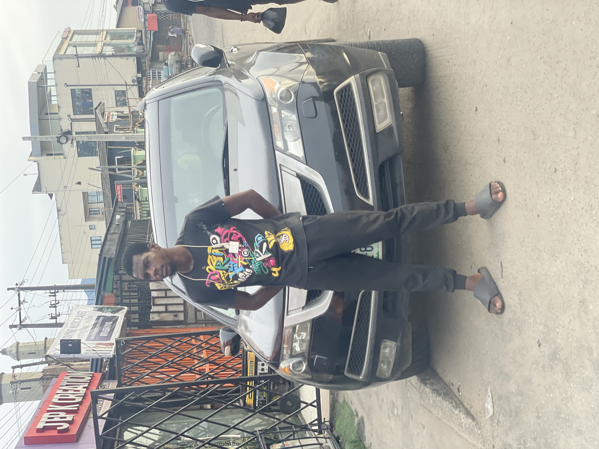 A young man stands in front of a dark SUV on a street in Ado, Nigeria. He wears a dark t-shirt with colorful graphics, dark pants, and gray sandals. The SUV is parked on the side of the road, partially blocking the view of the buildings behind it. A sign for "J.E.P.K Creation" is visible on the left. The street is paved and appears to be in a commercial area. Other buildings and street signs are visible in the background. A second person is partially visible above the SUV.