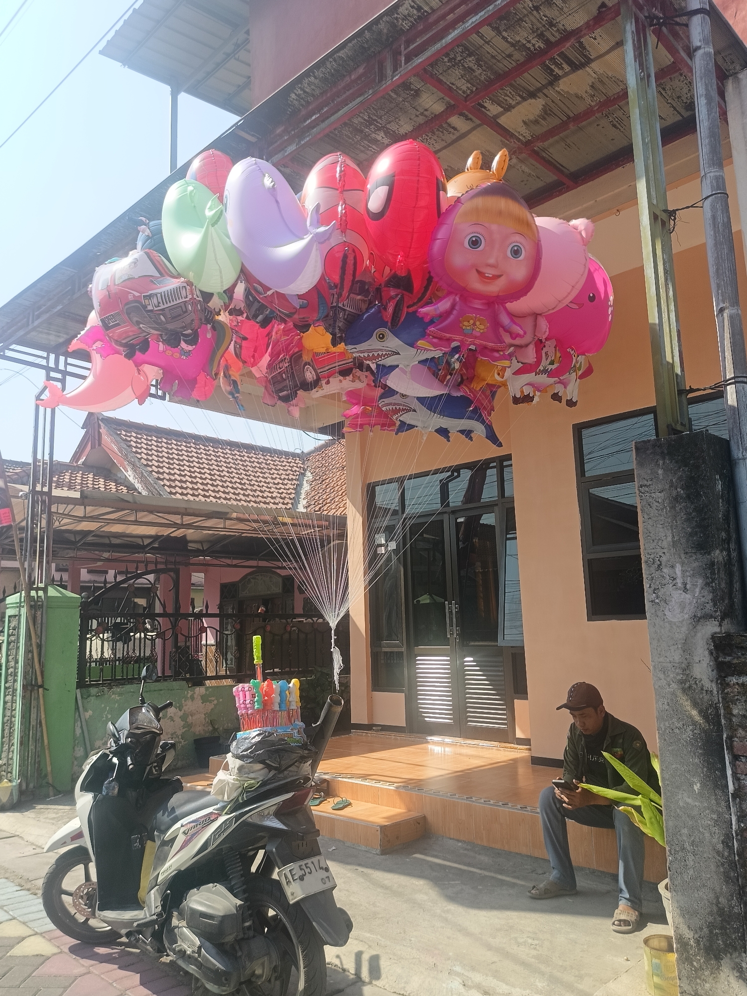 The image depicts a street scene in Gedangan, Indonesia, with a focus on a vendor's display of colorful balloons. A large cluster of Mylar balloons, featuring characters like Spider-Man, Masha and the Bear, and various animals and vehicles, is suspended from a canopy. In the foreground, a white and black motorcycle is parked, equipped with a basket containing more balloons. To the right, a man wearing a cap, dark jacket, and grey pants is seated on a ledge, looking at his mobile phone. The backdrop consists of a building with orange walls and dark-framed windows and doors, a tiled roof, and a fence. The ground is paved with tiles and concrete. The lighting suggests it is daytime.