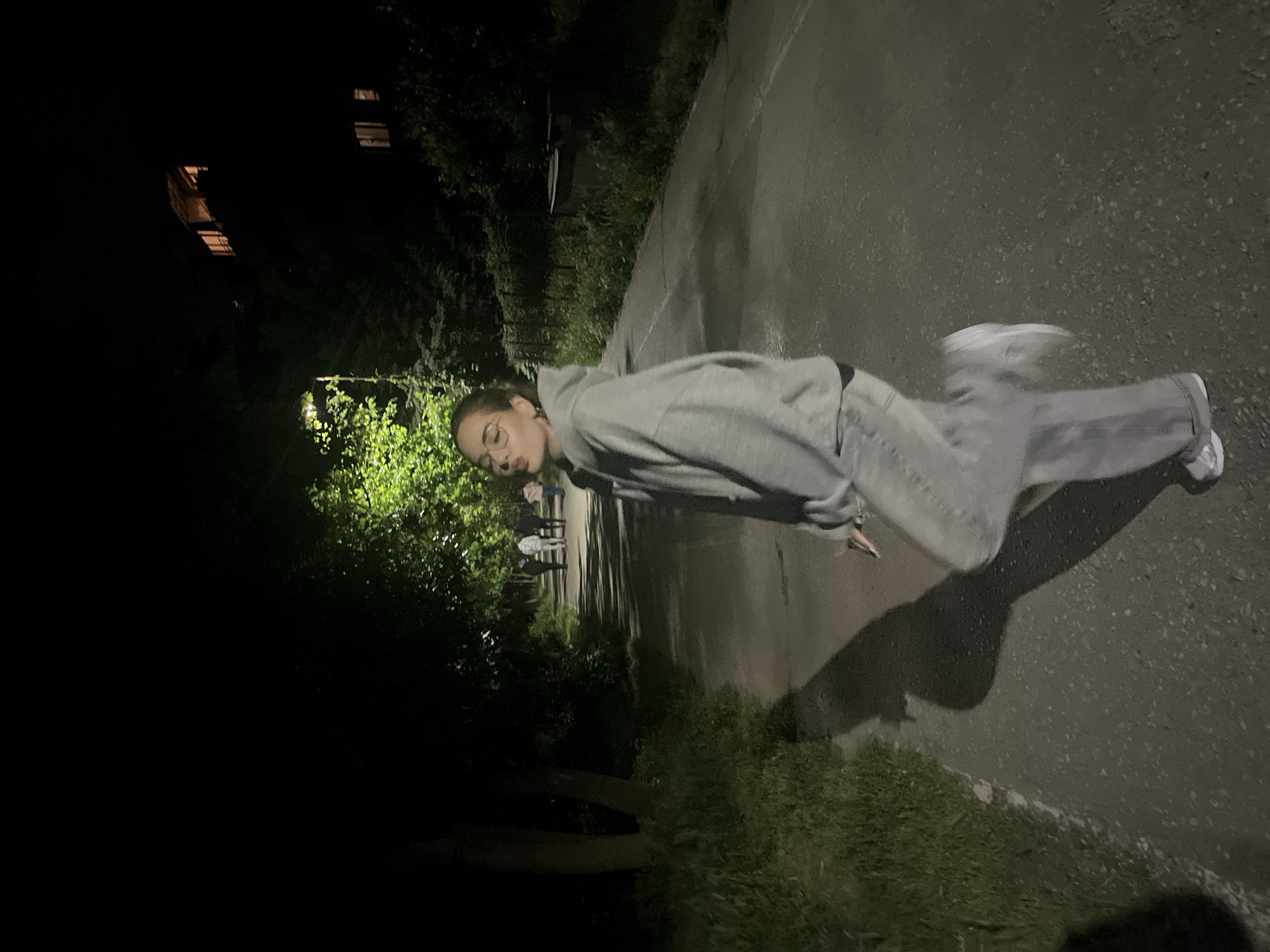 A medium shot depicts a woman on a dimly lit street at night. The woman is positioned on the right side of the frame, facing towards the left, and is dressed in a gray hoodie and matching sweatpants. Her head is turned slightly towards the camera with a slight pucker to her lips, possibly kissing towards the viewer. Her hair is pulled back.
The street surface appears to be asphalt, showing signs of wear. To the left of the woman, there is a strip of grass bordering the pavement, and beyond that, a darker area with trees and bushes. In the far distance, there's a faint indication of buildings, potentially illuminated by artificial light. Other people can be seen in the background, but are too far away to identify.
Based on the information provided, this image may be located in Blagoevgrad, Bulgaria, though this cannot be confirmed from the imagery alone.