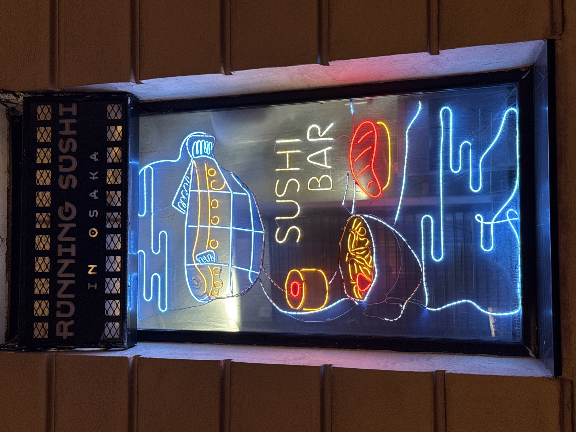 A nighttime view captures a window display of a sushi bar. The window is framed by a beige brick façade. To the left, a vertical sign reads "RUNNING SUSHI IN OSAKA" in capital letters. The window displays neon signs depicting sushi dishes and the words "SUSHI BAR" in a vertical arrangement. The location is not stated.