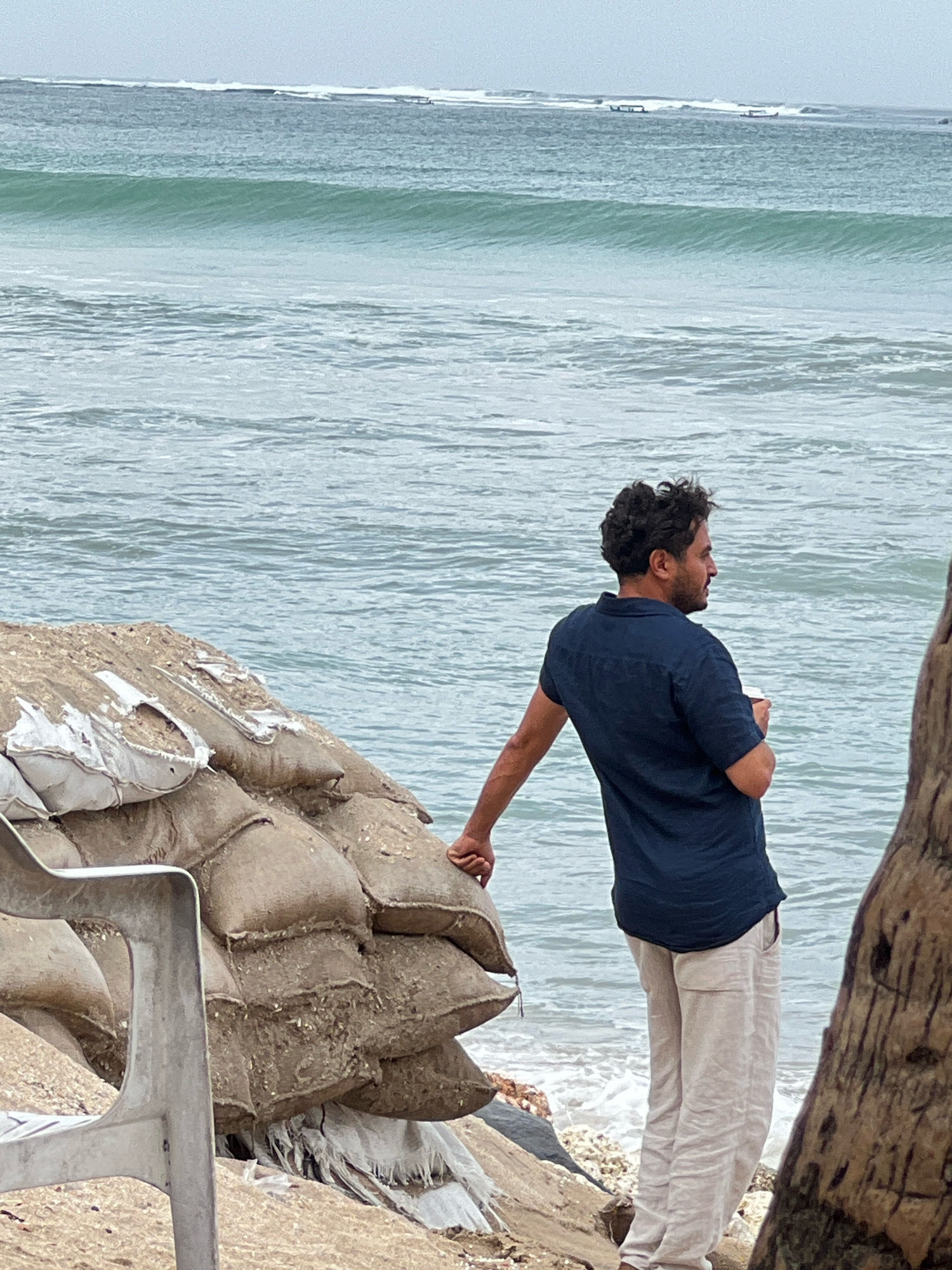 A man stands on a beach in Kuta, Indonesia, holding a white cup and looking out at the ocean. He is wearing a dark blue short-sleeved shirt and light-colored pants. In the foreground, there is a pile of sandbags, some of which are torn and frayed. A white plastic chair is partially visible on the left side of the frame. The ocean has moderate waves, and two small boats are visible in the distance. The sky is overcast.