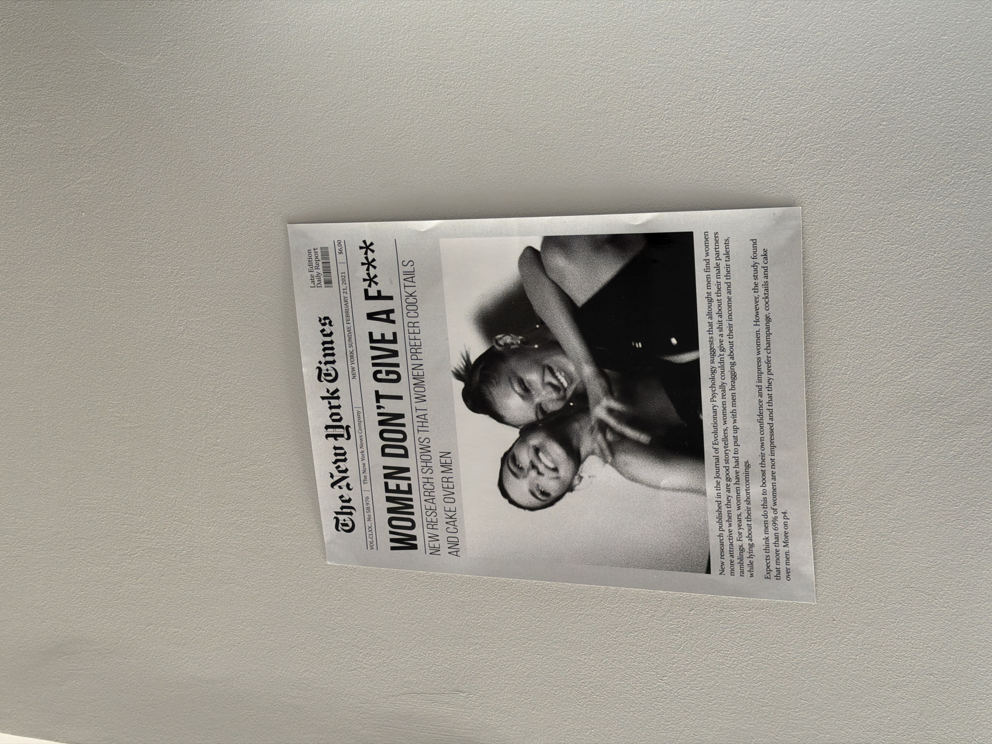A printed replica of The New York Times newspaper, dated Sunday, February 21, 2021, is displayed on a light gray, textured wall. The headline reads, "WOMEN DON'T GIVE A F*** NEW RESEARCH SHOWS THAT WOMEN PREFER COCKTAILS AND CAKE OVER MEN." Below the headline, a black and white photograph features two smiling women. The text on the newspaper discusses research suggesting women are more attracted to good storytellers and don't appreciate men bragging about their income or talents. It also states that most women are not impressed by men and prefer champagne, cocktails, and cake. The image is taken from a slight angle, with the newspaper appearing to be a poster or a framed print. The location is identified as Sirius, Russia, based on external context not present in the image itself.