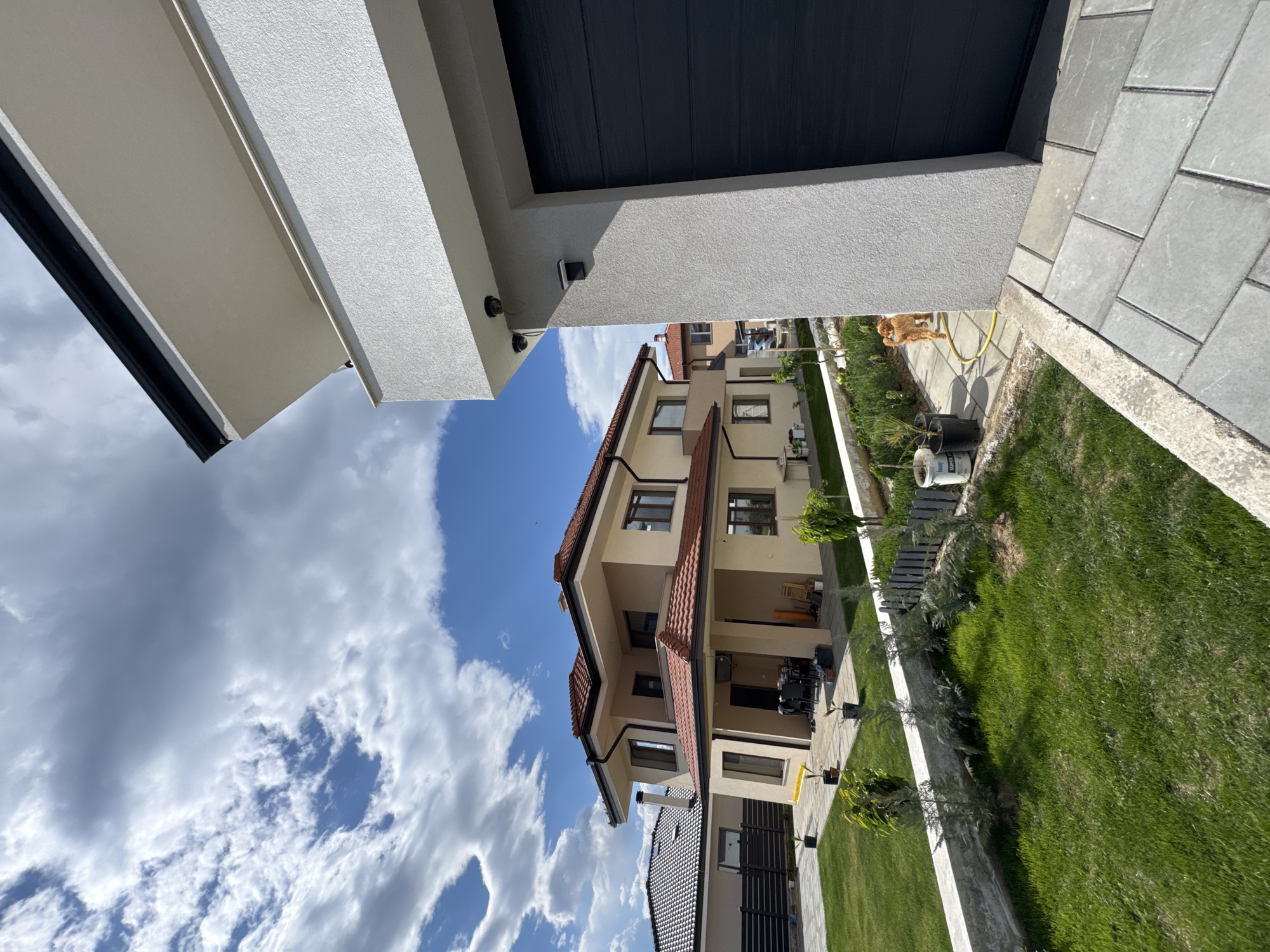 The image shows a two-story house with a red-tiled roof, tan exterior walls, and dark window frames. Adjacent to it is another building with a dark roof and light walls. The buildings are situated in a residential area with green lawns and trees. The sky is visible with blue patches and white clouds. The image is taken from a low angle, possibly from the ground level, and includes parts of structures immediately above, such as a white exterior wall and a dark-paneled ceiling. While the location is likely in a city in Bulgaria, and possibly Blagoevgrad, this cannot be definitively confirmed from the image alone.