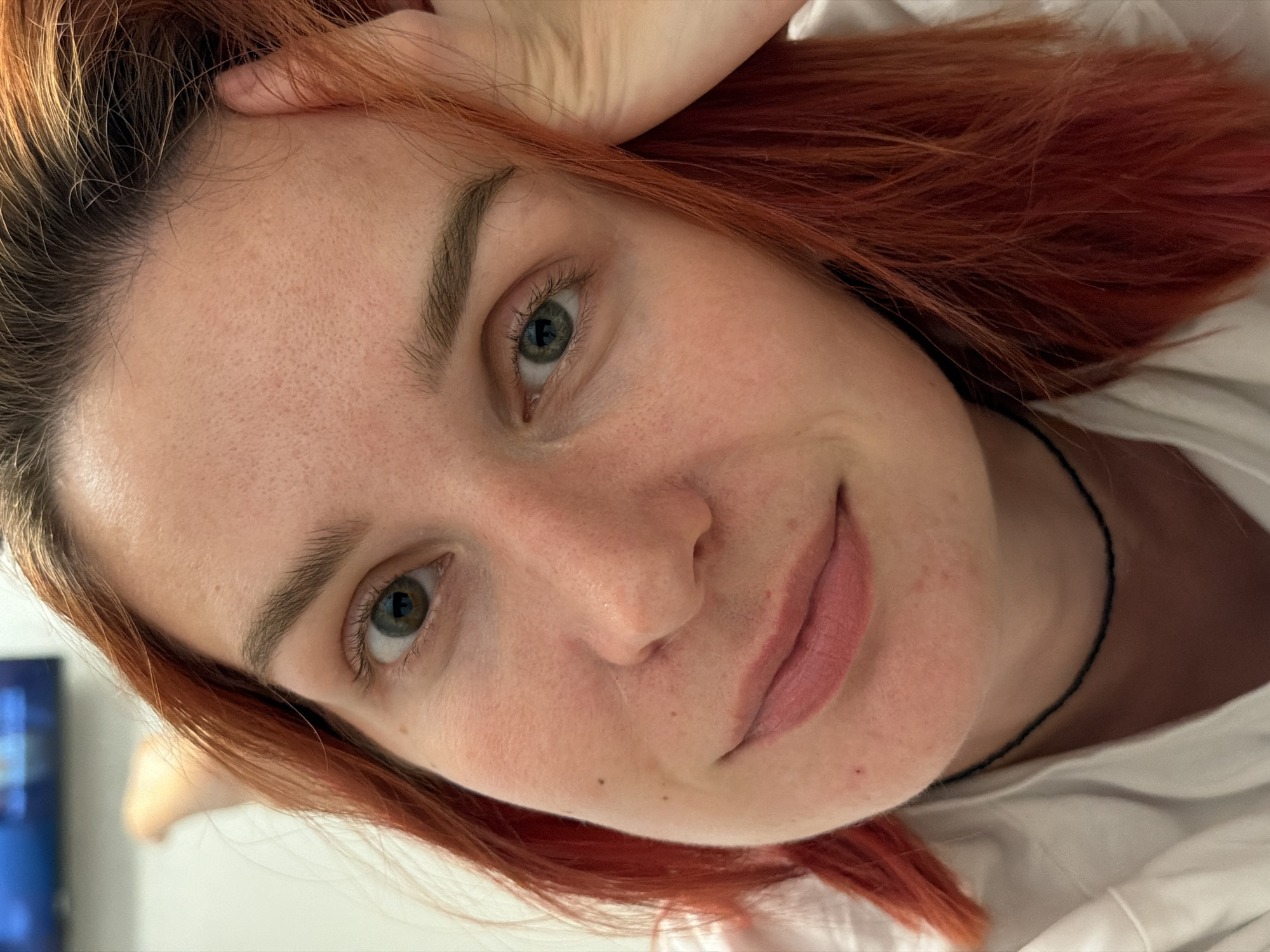 A close-up, slightly tilted, indoor portrait of a fair-skinned woman with red hair and green eyes. Her left hand rests on her forehead. She has light-colored eyebrows and pink lipstick. A thin black necklace is visible around her neck. She appears to be wearing a white shirt. A blurred television screen is visible in the background. There is no definitive indication of the location being Naberezhnye Chelny, Russia.