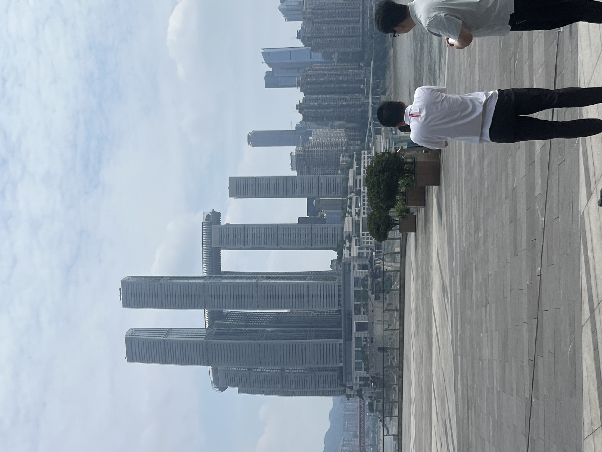 Two men stand on a paved plaza overlooking a city skyline. The man on the left, wearing a white polo shirt and black pants, holds a phone up to take a photo. The man on the right, wearing a light green polo shirt and black pants, holds a phone in his right hand. In the background, a large modern complex of connected skyscrapers dominates the view. A wide river is visible in the middle ground, with a red bridge crossing it in the distance. The sky is overcast with scattered clouds. The location is identified as Nan'an District, China.