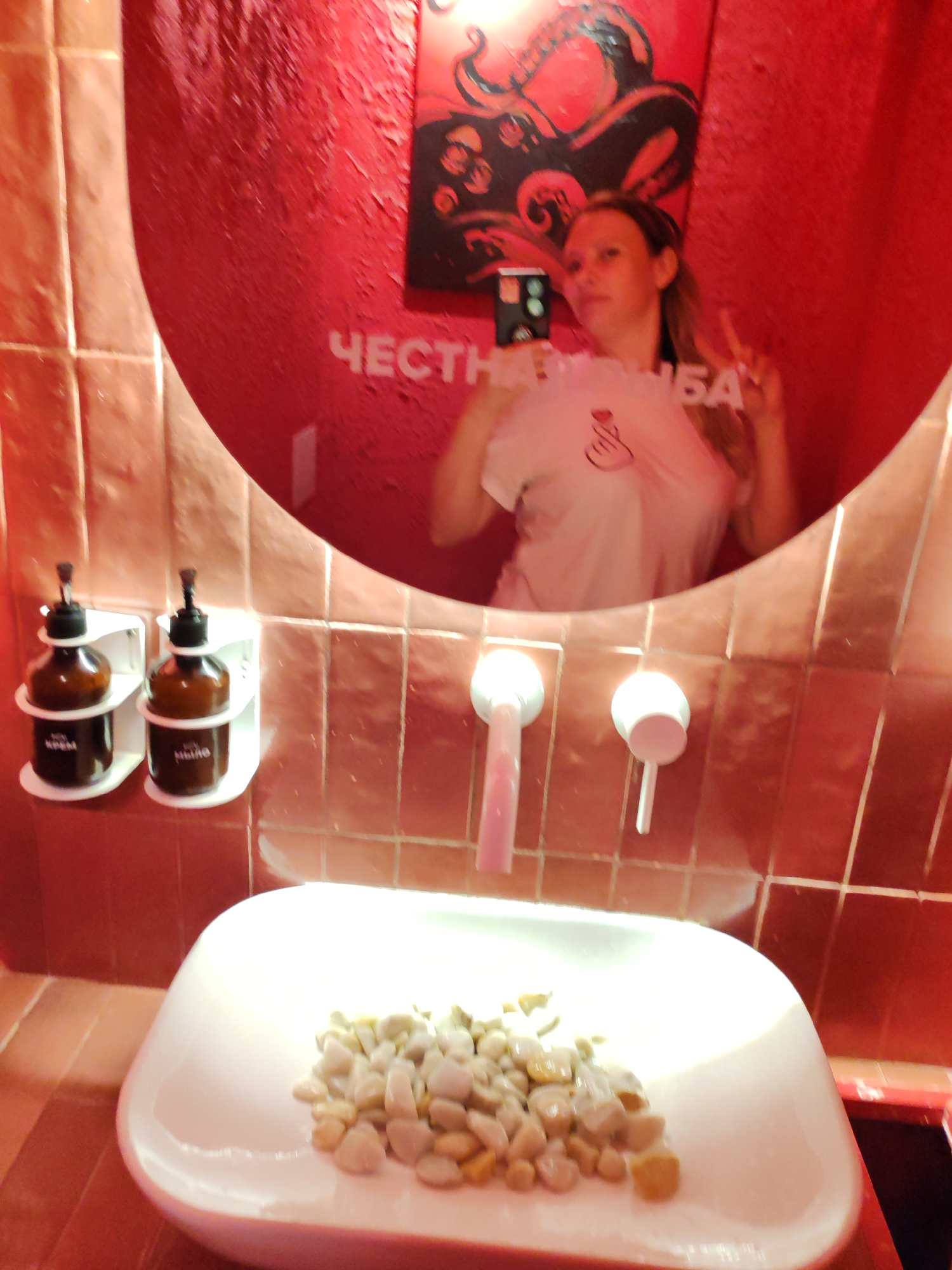A young woman takes a mirror selfie in a bathroom with red tiled walls and a large, oval mirror. She is wearing a light-colored t-shirt with a pink graphic of a hand making a heart shape. In the reflection, text on the wall above her reads "ЧЕСТН" and "ЬБА". A black and red octopus graphic is visible on the wall behind her.
In the foreground, a white sink is filled with small, light-colored stones. Two brown soap dispensers are mounted on the wall to the left of the sink. The sink area has two white faucets. The overall impression is a modern, stylish bathroom with a playful artistic touch. The lighting suggests it is likely daytime, but the artificial lighting in the bathroom also contributes to the ambiance. The woman appears casual and relaxed, posing for a photo. The location is specified as Moscow, Russia.
