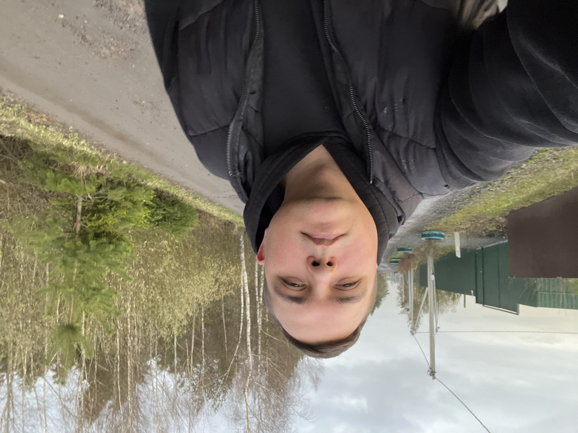 A selfie captures a young man from the chest up, looking directly at the camera with a neutral expression. He is dressed in a dark, likely black, puffer jacket worn over a dark hooded sweatshirt.
The background reveals a rural or semi-rural landscape. To the man's left, a dirt or gravel road runs parallel to a strip of green grass and small evergreen saplings. Further back, a mix of bare-branched deciduous trees, notably white-barked birches, and some evergreen trees form a forest line under an overcast sky. To his right, the road continues, passing a dark green building, possibly a shed or workshop, and several utility poles with overhead wires. A few small blue-capped posts are also visible near the road on the right.
The scene suggests an outdoor setting on an overcast day, indicating cooler weather consistent with the man's attire. The ground appears somewhat damp. The activity is the man taking a selfie. No specific emotions are conveyed, and no visible text is present. The setting is consistent with a countryside or village environment in Razdolye, Russia.
