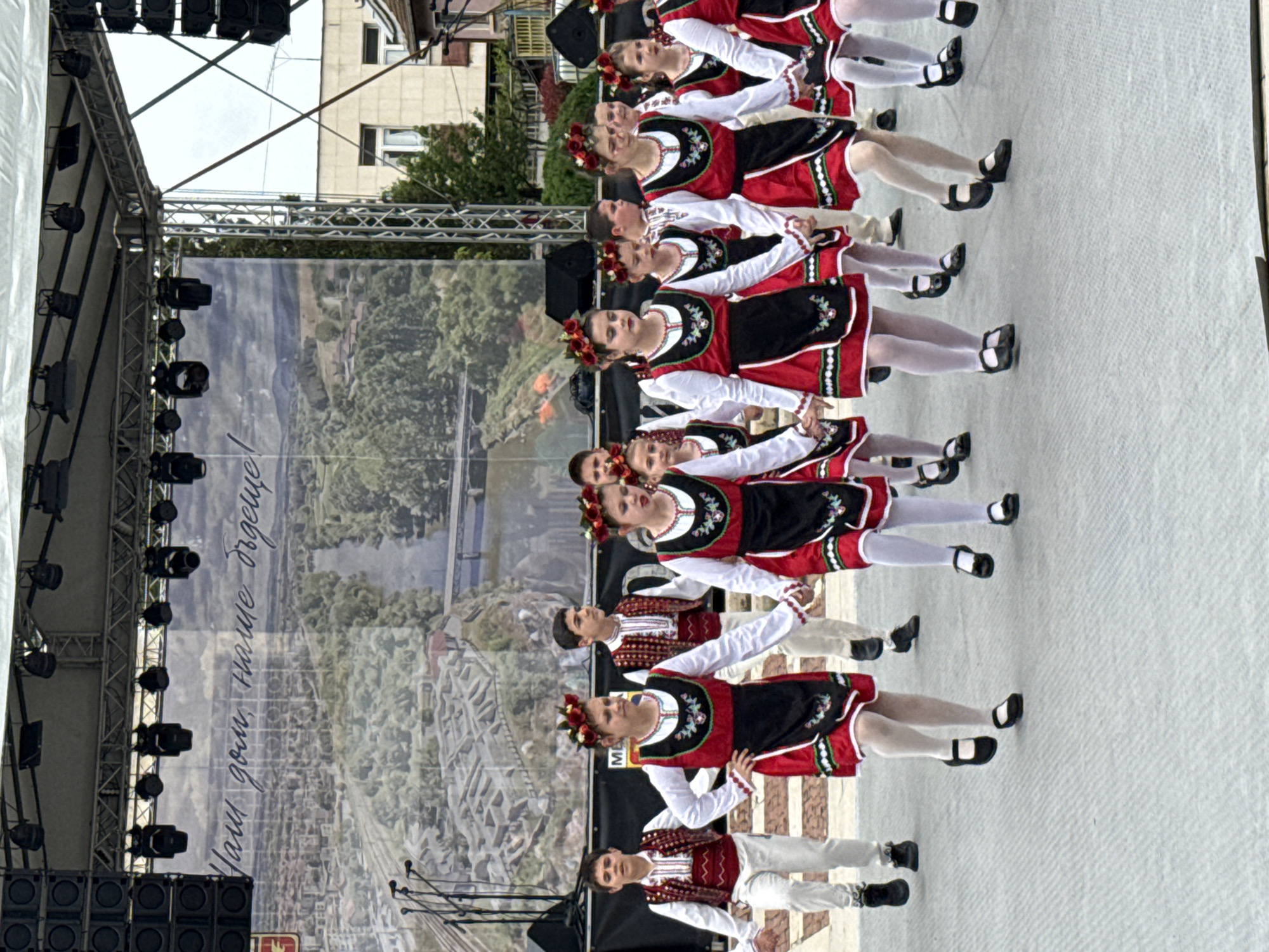 A line of ten dancers in traditional Bulgarian folk costumes are performing on an outdoor stage in Mezdra, Bulgaria. The dancers, mostly women, are wearing red and black dresses with white sleeves and floral headpieces. The sole male dancer is dressed in white pants and a red and white embroidered vest. All are wearing dark shoes with buckles. They are holding hands and performing a coordinated dance, likely a traditional Bulgarian folk dance. The backdrop of the stage features a large banner displaying a scenic view of Mezdra, likely its architectural landmarks, featuring a river and a historical landscape. The stage itself is made of a white surface. Stage lighting and speakers are visible on the left. A building is partly visible in the background. The weather appears overcast.