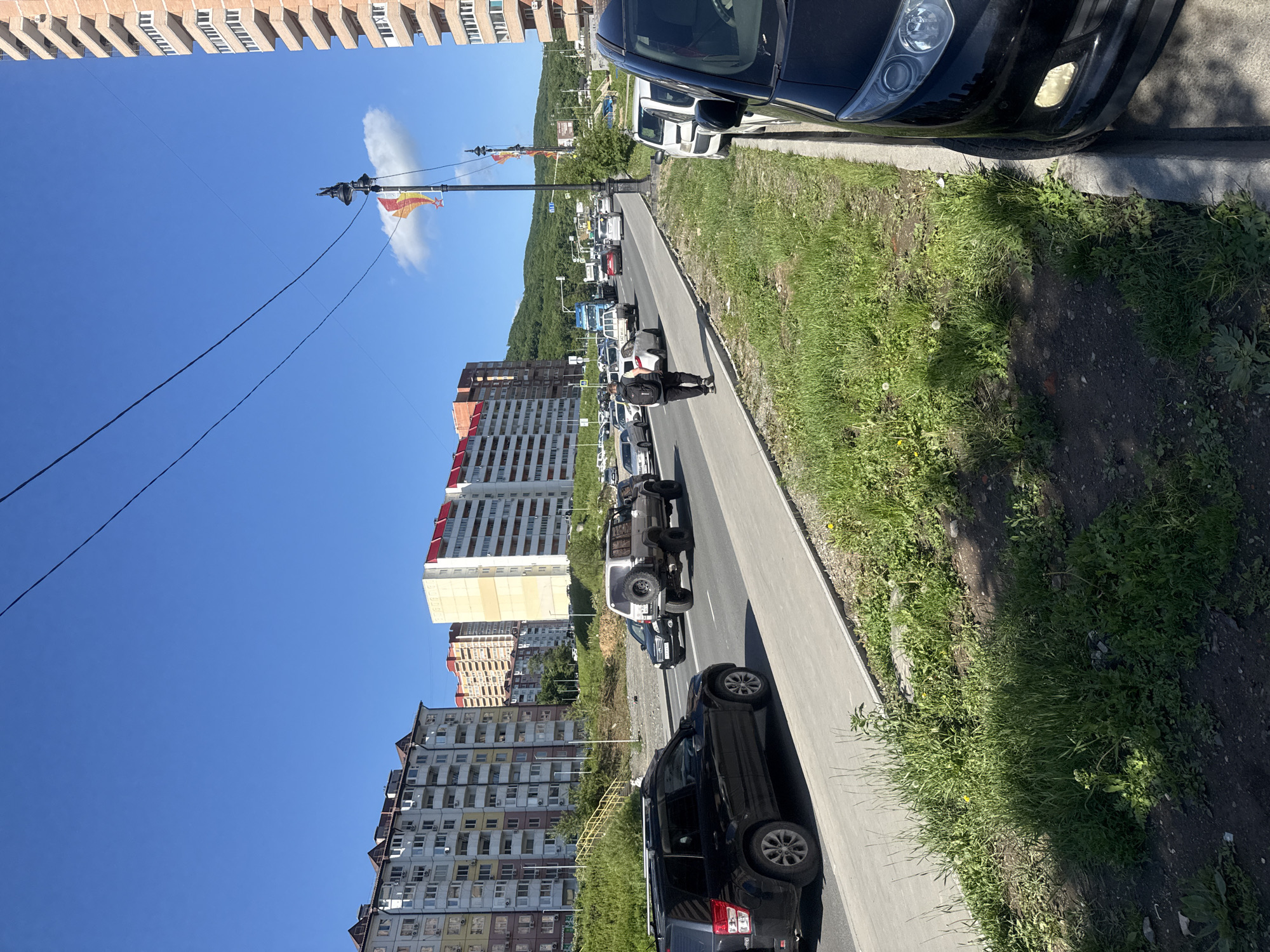 The image depicts a street scene in Vladivostok, Russia, under a clear blue sky. Multiple apartment buildings, characterized by their multi-story structures and varied architectural styles, line the background. Some buildings feature red-tiled roofs, while others have flat tops. In the foreground, a paved road is occupied by several vehicles, including a black SUV, a silver off-road vehicle, and other cars. A pedestrian, carrying a backpack, walks along the sidewalk bordering the road. Green grass and vegetation grow on the embankment next to the sidewalk. A lamppost with decorative flags is visible on the right side of the road. The scene appears to be a moderately busy urban area, possibly during the daytime.