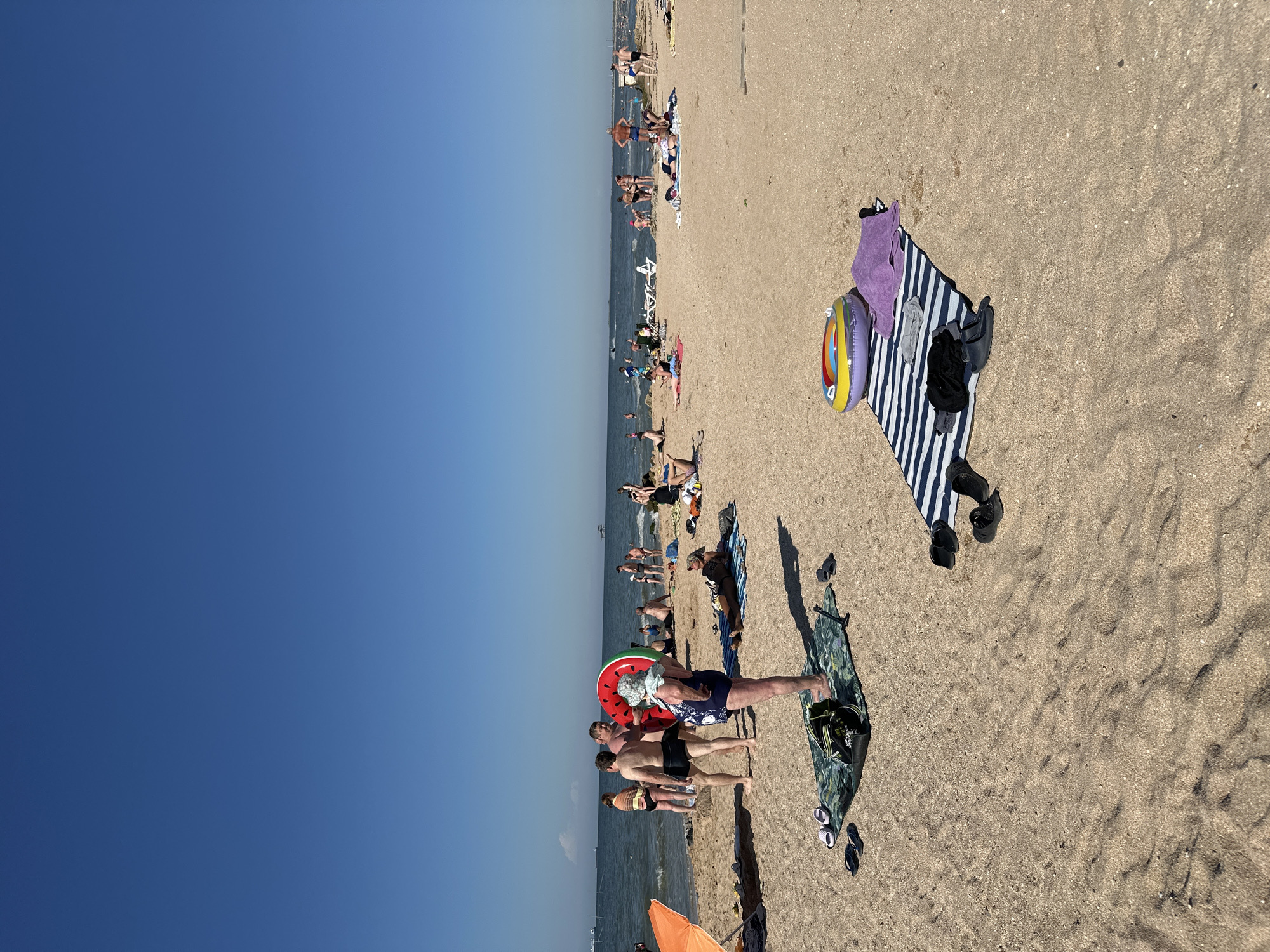 A sunny day at Golubitskaya, Russia's Black Sea beach is captured from a low angle perspective. The sky is a clear, vibrant blue with no clouds. The beach is populated with numerous individuals enjoying the warm weather. In the foreground, a woman wearing a blue and white patterned swimsuit and a light-colored sun hat stands on a green and black patterned beach mat. Next to her, a person is partially visible, leaning over to pick up an item from the mat. Several pairs of flip-flops are scattered on the sand around the mat.
Further back, closer to the water's edge, a group of people are gathered. Two men in swim trunks are talking, one of whom is holding a large, inflatable watermelon. Behind them, more people are visible, some standing and others relaxing on the sand or on towels. The sea is a calm blue, with small waves lapping at the shore. In the distance, various small boats and what appear to be oil rigs are scattered across the horizon. Scattered throughout the beach are towels, beach mats, and inflatable toys, indicating a typical day of recreation. The sand is a light brown and appears to be a mixture of fine grains and small shells. The overall atmosphere is one of leisure and enjoyment under the summer sun.