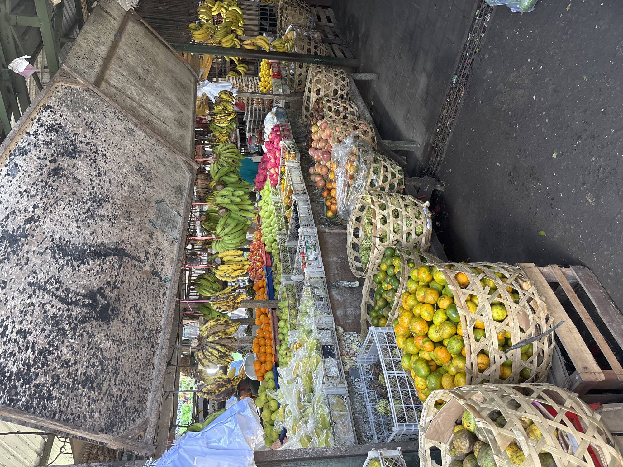 The image shows a vibrant fruit market in Ungasan, Indonesia. The scene is an outdoor stall under a weathered roof. Various fruits, including bananas, mangoes, oranges, star fruit, dragon fruit, and salak are displayed. Bananas hang from the roof of the stall, while other fruits are arranged in baskets, wooden crates, and plastic containers on shelves. The ground is paved and appears damp. A person is partially visible near the bottom of the frame. The atmosphere is characteristic of a local Indonesian marketplace.