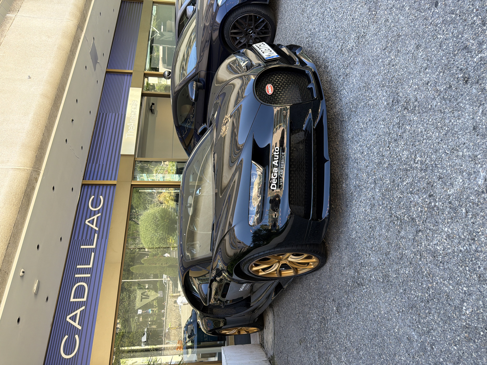 The image shows a black Bugatti Chiron parked on an asphalt street in front of a building with "CADILLAC" and "The Arsenal" signs. The building has a modern facade with glass windows and blue striped accents. The Bugatti has gold wheels and a sticker on its front fender that reads "DeGa Auto VIP Car Service." To the right of the Bugatti, a dark-colored Mercedes-Benz coupe is parked. The environment appears to be an urban setting, possibly a dealership or a luxury car service center, given the presence of high-end vehicles and the branding. The lighting suggests it is daytime, with sunlight casting shadows.