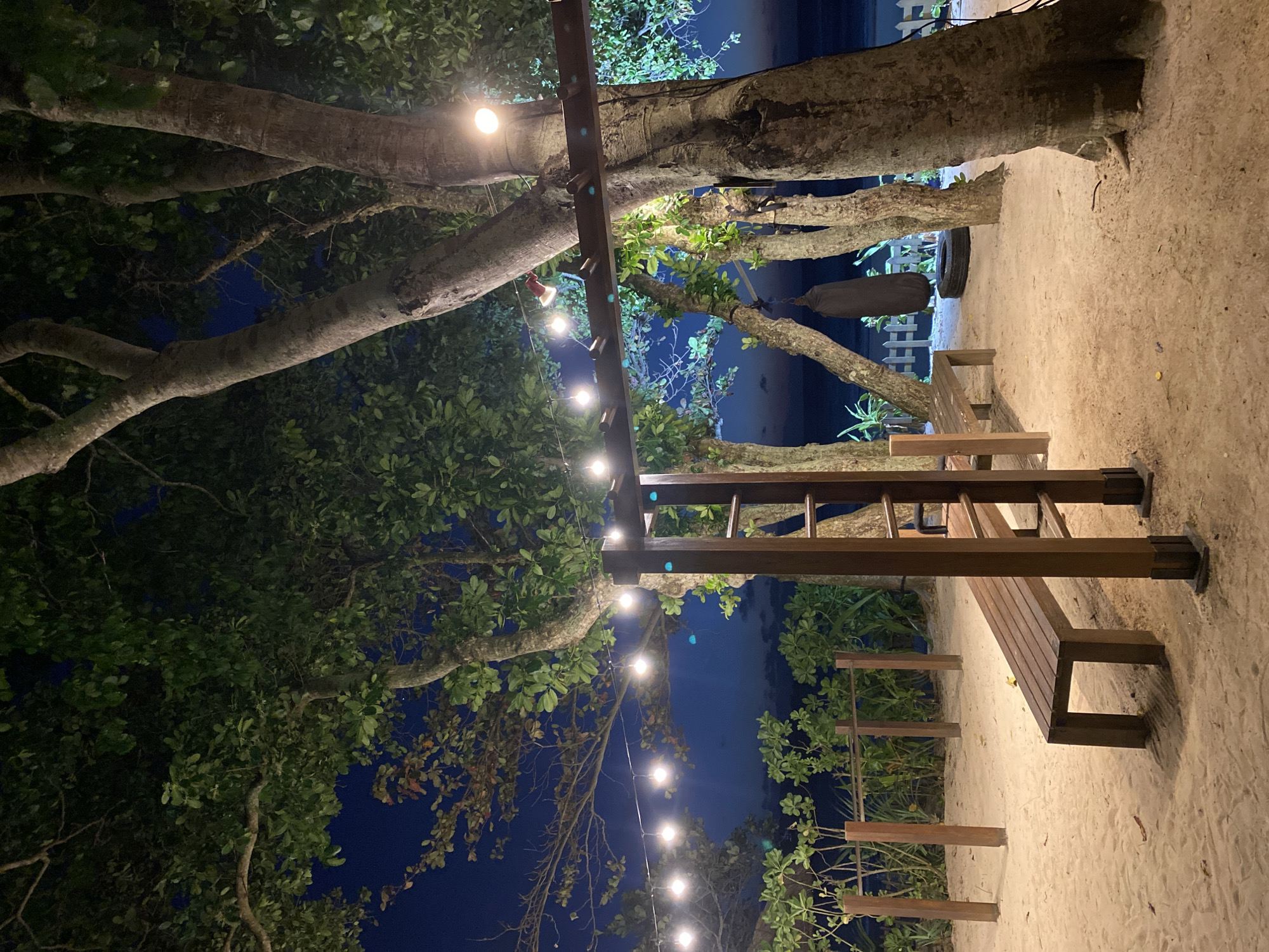 The image captures an illuminated outdoor scene at night, set in what appears to be a tropical environment, consistent with Kuta Selatan, Indonesia. Large trees with thick, textured trunks and abundant green foliage dominate the left and upper portions of the frame. A dark, deep blue night sky is visible through the gaps in the tree canopy.
The area is softly lit by strings of warm, glowing light bulbs, suspended horizontally across the scene. These lights appear to be strung from a sturdy wooden beam, which is part of a larger wooden structure. This structure, visible on the right, features upright posts and horizontal slats, resembling a rustic pergola or decorative screen. A simple wooden bench with a slatted design is situated on the light-colored sandy ground towards the mid-right of the image.
The ground is entirely covered in sand, suggesting a beachside or sandy garden location. In the background, a low white picket fence is partially visible behind some tree trunks and plants. Beyond the fence, a faint dark line against the horizon suggests the presence of water, likely the ocean, under the moonless night sky. Additional green plants and bushes are scattered around the scene, enhancing the natural setting. The artificial lighting creates a warm, inviting, and tranquil atmosphere. No people or visible text are present in the image.