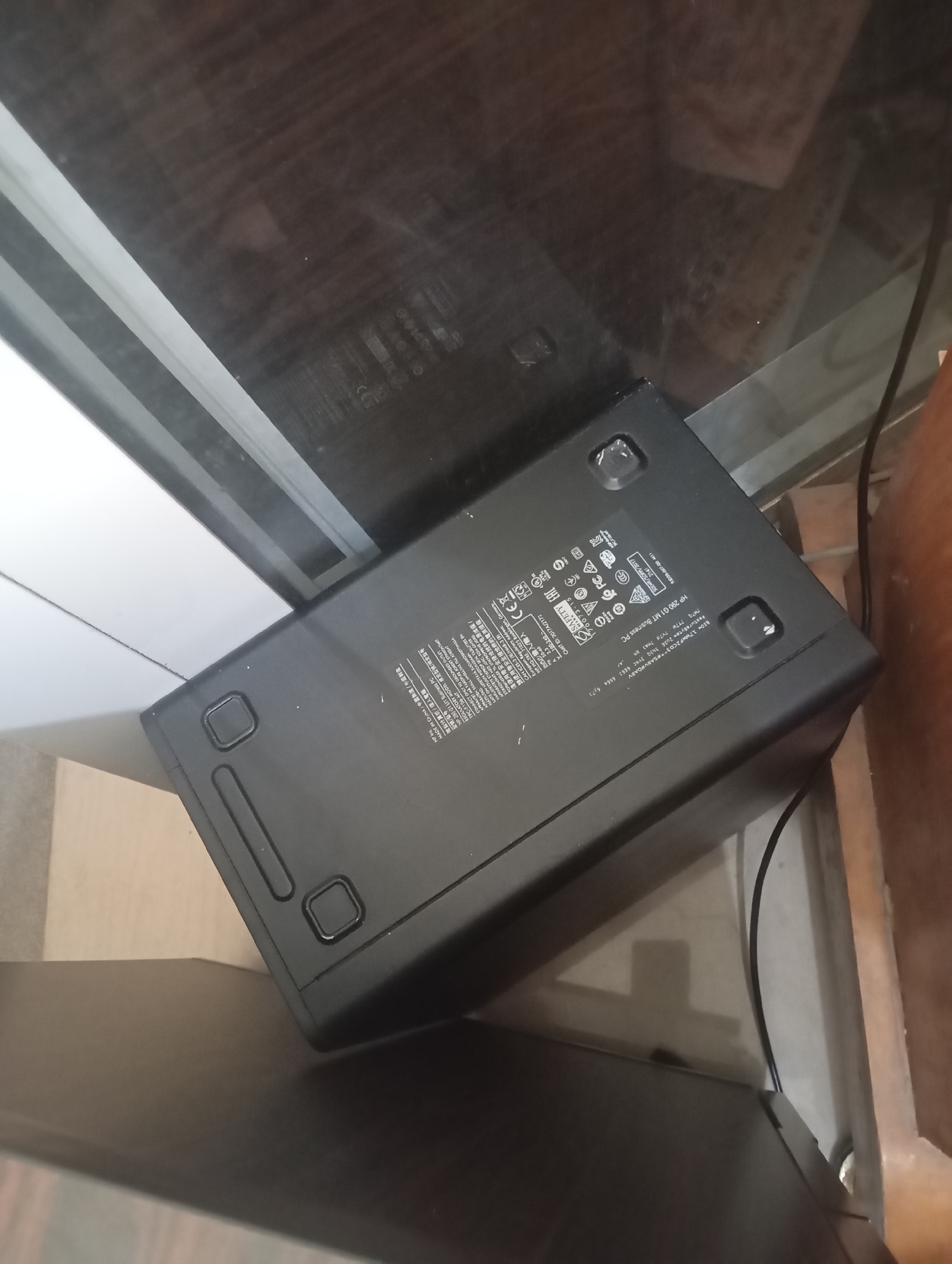 The image shows the bottom of a black HP 280 G1 MT BUSINESS PC, likely a desktop computer tower. It is lying on its side on a dark, reflective surface, possibly a glass tabletop. Several labels are visible on the underside of the PC, including regulatory markings, model information, and country of origin (MADE IN CHINA). There are also rubber feet on the bottom of the PC, two of which are clearly visible. A black power cable is also partially visible extending from the right side of the PC. The background is dark and out of focus, with hints of furniture and a wall. There is no indication of the specific location within Jos, Nigeria, nor are there any people or discernible activity depicted in the image. The scene appears to be an indoor setting, likely a room in a home or office.