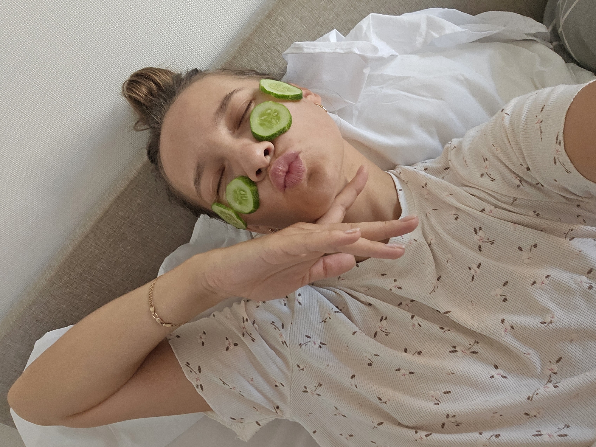 The image shows a young woman lying on a bed with her eyes closed and cucumber slices on her eyelids and under her eyes. She is puckering her lips and holding up one hand in a peace sign. The woman is wearing a light-colored, ribbed t-shirt with a small floral pattern. She has a gold bracelet on her wrist. The background is a white sheet and a beige headboard. The image appears to be a selfie taken from a high angle. The location of Chaikovsky, Russia is not visible in the image.