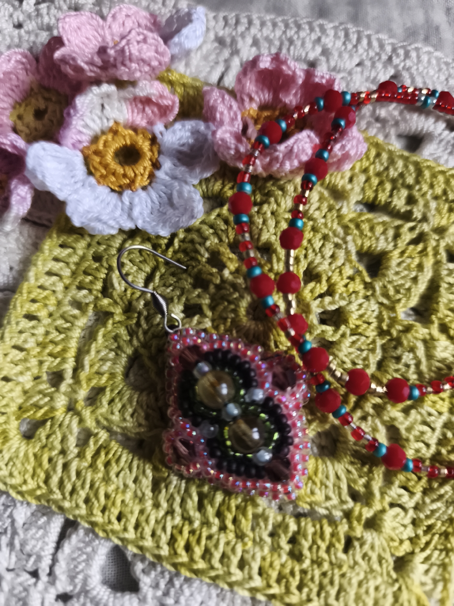 The image displays a close-up view of handmade crafts, specifically crocheted flowers and beaded jewelry, laid out on crocheted fabric. The crocheted flowers are in shades of pink, white, and yellow, with intricate petal designs. A beaded necklace, featuring red, teal, and gold beads, is draped across the scene, and a single beaded earring with a square pendant is visible. The background consists of yellow and white crocheted textiles, creating a textured surface for the display. There is no indication of location in the city of Saint-Denis, France. The objects are stationary and arranged for aesthetic presentation.