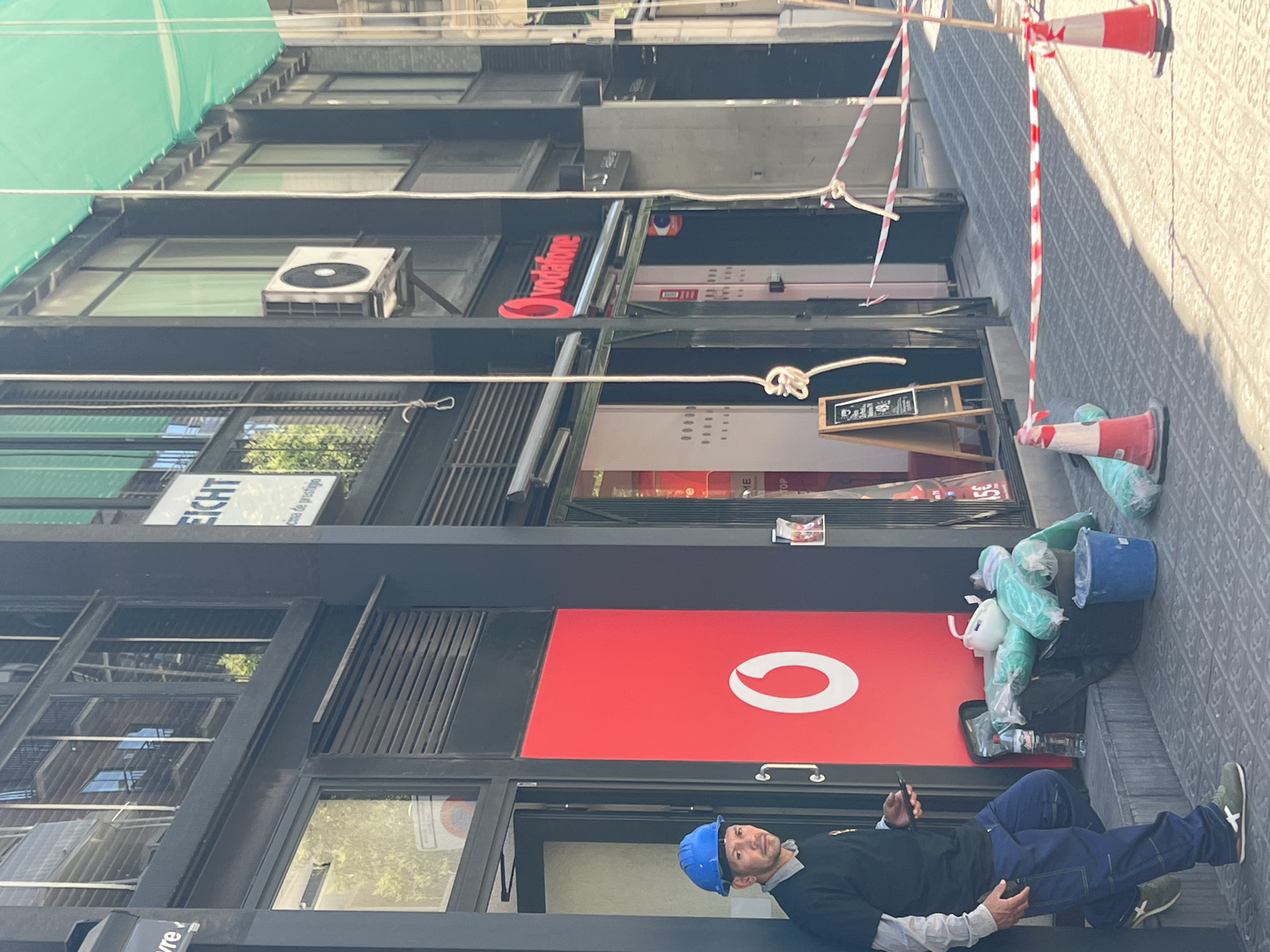 The image captures a street-level view of a commercial building on a sunny day in Barcelona, Spain. A man wearing a blue hard hat, a dark t-shirt, and blue work pants stands near the entrance of a store with a large red facade featuring a prominent white Vodafone logo. He is holding a smartphone in his left hand. To his right, a black bag, a blue bucket, and several large plastic-wrapped items are placed on the ground, along with a partially obscured water bottle. A wooden A-frame sign with chalk writing stands in front of a store entrance further down the street. Red and white striped caution tape, supported by orange traffic cones, is set up across the sidewalk, suggesting ongoing work or a restricted area. The building has dark framing around its windows, and an air conditioning unit is visible mounted on the exterior wall. Reflections of buildings and trees can be seen in the glass windows of the upper floors.