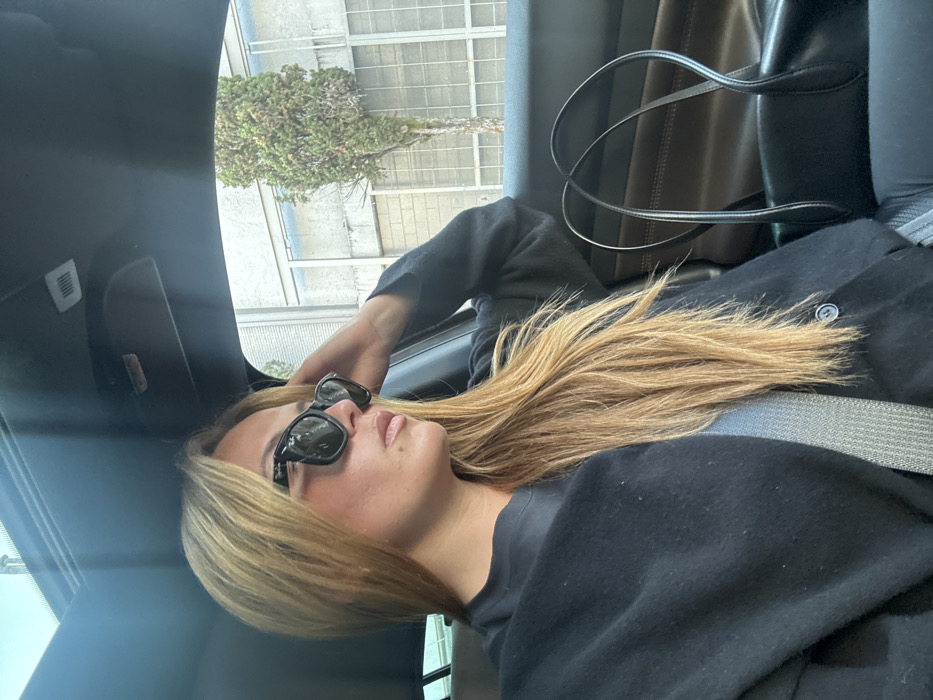 A woman with long blonde hair, wearing sunglasses and a black sweater, lies back in what appears to be the passenger seat of a car. She is wearing a seatbelt. Sunlight streams in from the window, illuminating her face. Outside, a building with green foliage can be seen through the sunroof. A black handbag hangs behind her.