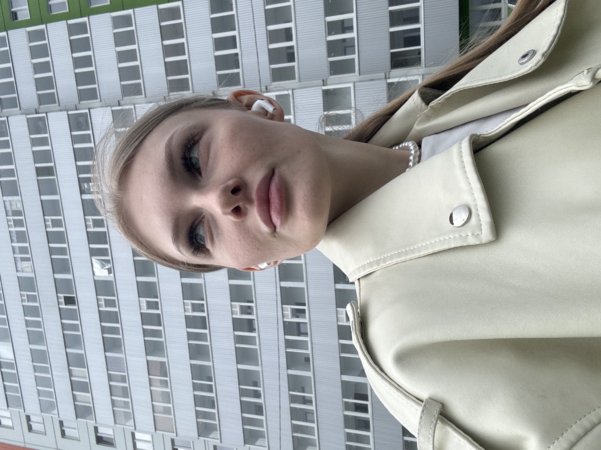 A medium close-up, portrait-oriented photograph features a young woman with fair skin, light-colored hair, and blue/green eyes, looking slightly away from the camera. The image is rotated clockwise, placing the building in the background at an angle.
The woman is wearing a light beige or cream-colored jacket, likely made of leather or faux leather, with silver snap buttons visible on the collar and lapel. A white pearl or bead necklace is visible around her neck, peeking out from under her jacket. She has white wireless earbuds (AirPods) in both ears, suggesting she is listening to audio. Her expression is neutral and composed.
In the background, a tall, multi-story building dominates the scene. Its facade consists of numerous rectangular gray/silver panels and evenly spaced, large rectangular windows, creating a repeating pattern. A section of the building's upper part is painted green. The building's design suggests a modern apartment or office complex in an urban or suburban setting. The lighting is diffused and natural, indicating daytime with soft, even illumination. There is no visible text in the image. The location context provided is Кондратово, Russia.