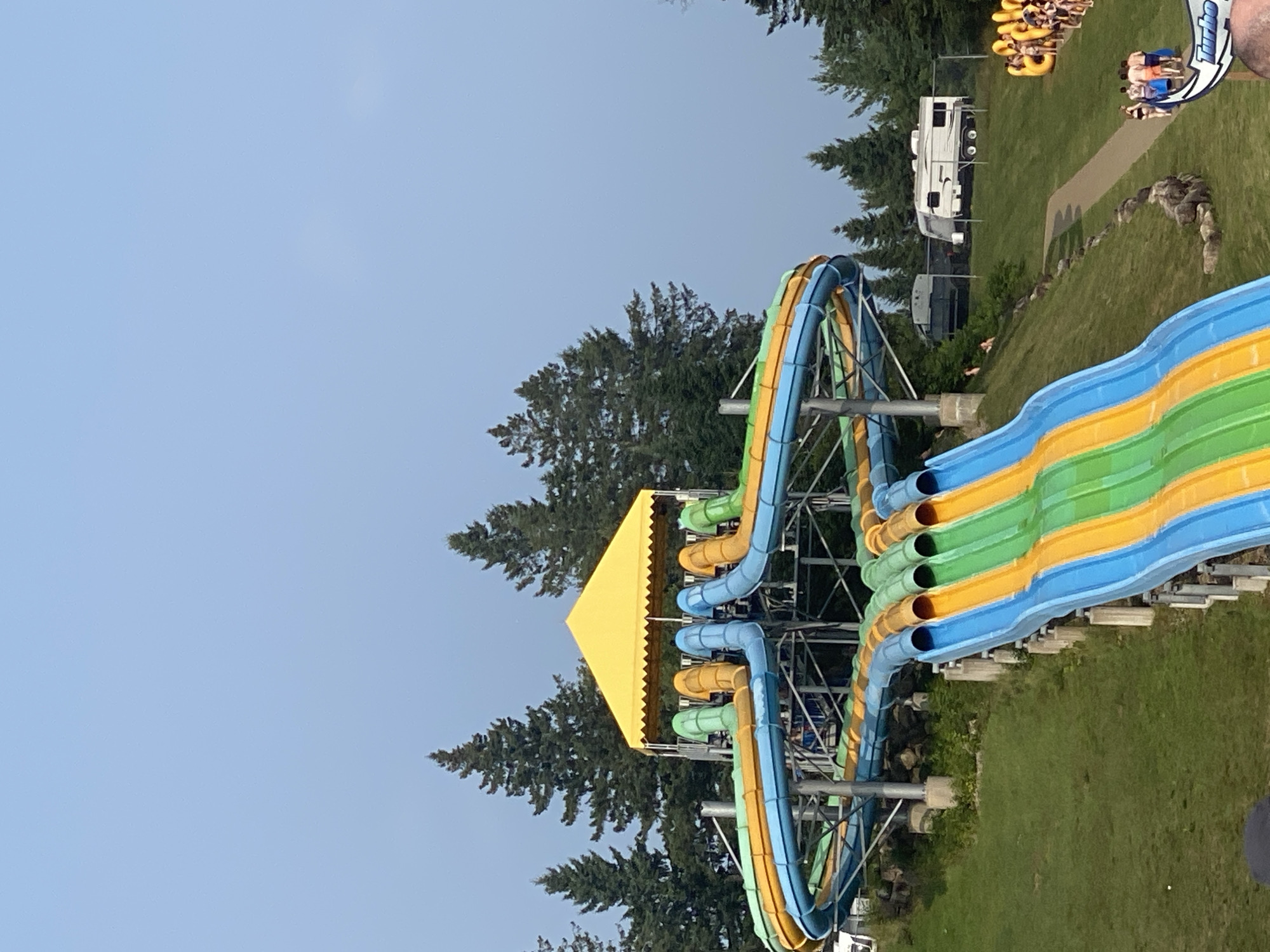 This image captures a scene at the Bora Parc water park, located in the city of Saint-Gabriel-de-Valcartier, Canada. In the foreground, a multi-lane water slide complex dominates the view. The slides are brightly colored in segments of blue, orange, and green, with a yellow canopy overhead at the top. The structure is supported by metal poles and concrete bases, situated on a grassy, sloped hill. Trees, primarily evergreen, form a backdrop behind the water slides.
Further into the scene, a paved pathway winds down the hill, leading towards a group of people and a line of yellow inner tubes. A white recreational vehicle (RV) is parked in the background, behind a fence, alongside another, smaller RV or trailer. The sky is a pale blue, suggesting a partly cloudy day. Several individuals are visible, some walking along the path, others queuing for the slides or inner tubes, and a few are seen on the grassy slopes. The overall impression is a recreational environment on a summer day, with the water park's attractions clearly visible.