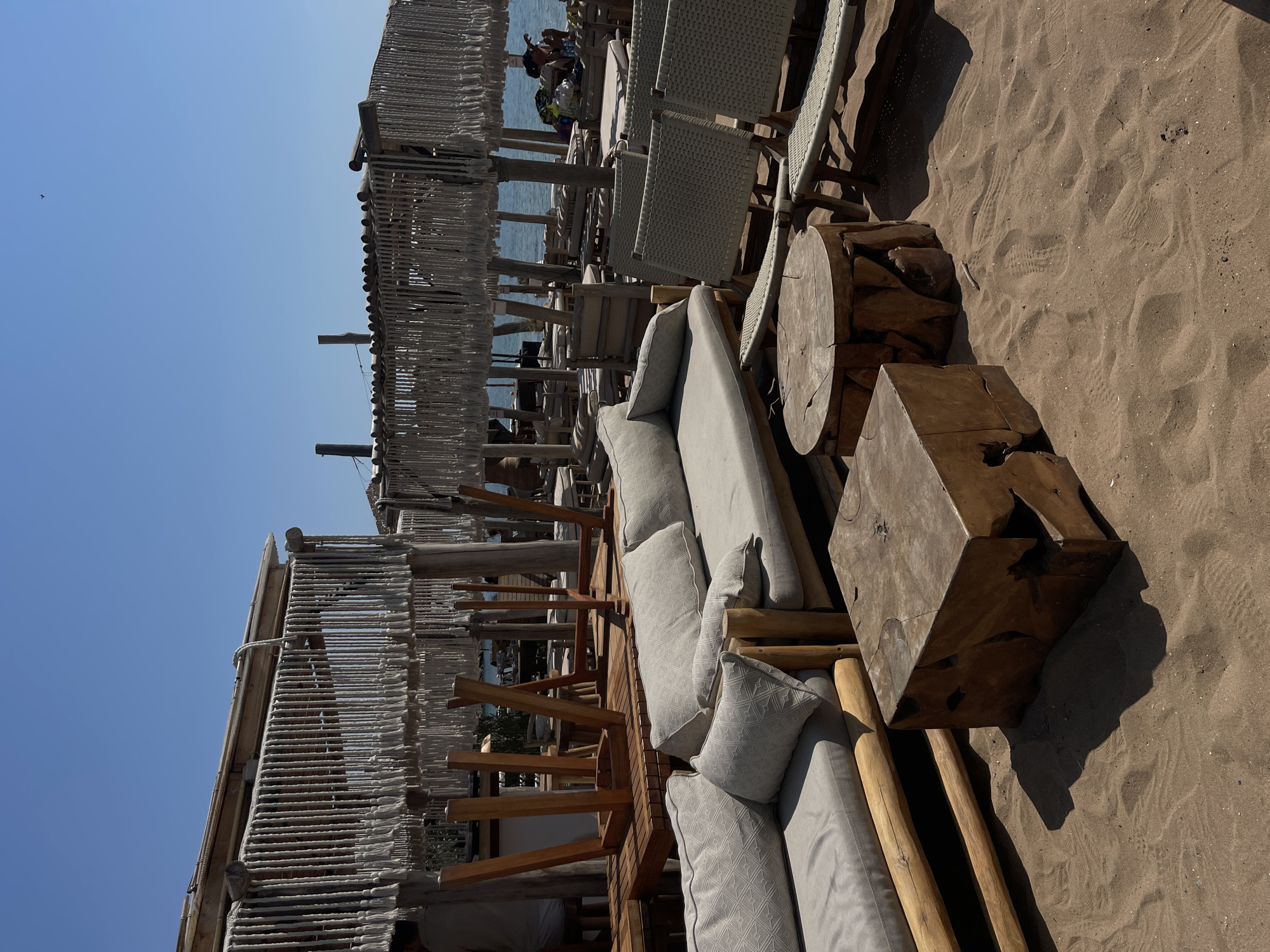 The image depicts a beachside seating area at a resort in Chernomorets, Bulgaria. The scene is set on a sandy beach with the sea visible in the background. In the foreground, there are several pieces of outdoor furniture made of natural wood and light-colored fabric. These include a large sectional sofa with multiple cushions and a low rectangular coffee table with a rustic, unfinished wood surface. To the right of the sofa, there are two woven chairs and a cylindrical side table also made of wood. Further back, there are more tables and chairs, some with cushions, and lounge chairs arranged under thatched roofs supported by wooden posts. The structures are decorated with rope detailing. In the mid-ground, there are several wooden tables with chairs stacked upside down on top of them, suggesting the area may be closing or preparing for the day. The sky is clear and blue. A few people are visible in the background, some seated on lounge chairs near the water.