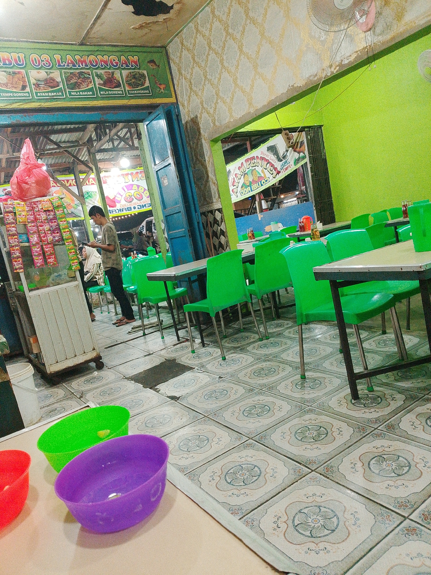 The image depicts a street-side food stall in Bandar Setia, Indonesia, possibly a restaurant specializing in Lamongan cuisine. A green banner with "BU 03 LAMONGAN" is prominently displayed at the top, featuring images of food items such as fried tempeh, grilled chicken, grilled fish, and stir-fried greens. A man in a grey t-shirt and black pants stands near a cart filled with packaged goods, looking at his phone. To his left, a woman in a headscarf is seated. The stall is furnished with brightly colored green plastic chairs and tables. The floor is tiled with a repeating floral pattern. In the foreground, three plastic bowls – red, green, and purple – are visible on a table. The stall appears to be open to the street, with various food items and beverages displayed on shelves. The overall atmosphere suggests a casual dining establishment catering to local patrons.